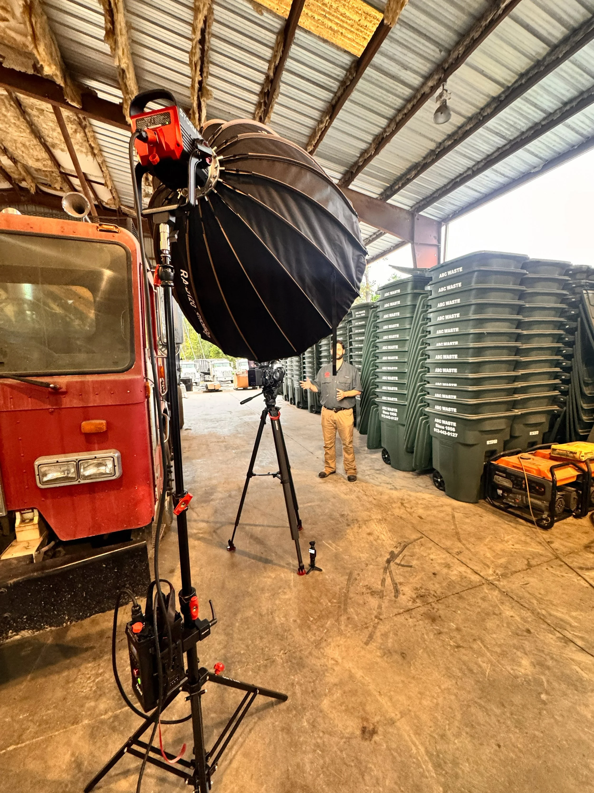 A large photography lighting setup with a black umbrella reflector on a stand, positioned near a red vintage vehicle inside a warehouse. Several stacks of green and black waste bins with white labels are along the wall, and a person is standing near 