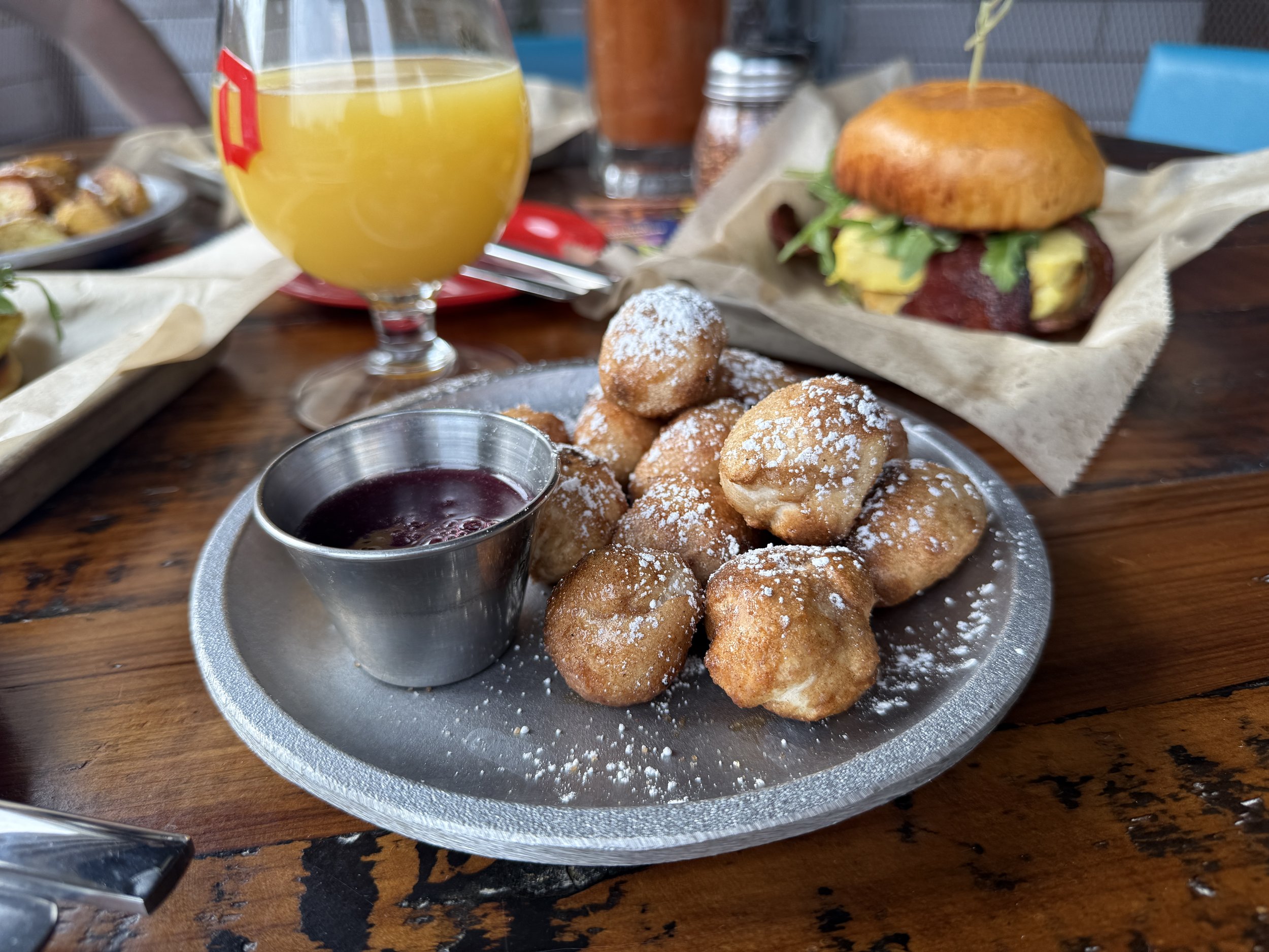 Fried dough balls with powdered sugar served with berry syrup, a breakfast sandwich with lettuce, bacon, and a biscuit, and a glass of orange juice on a wooden table.