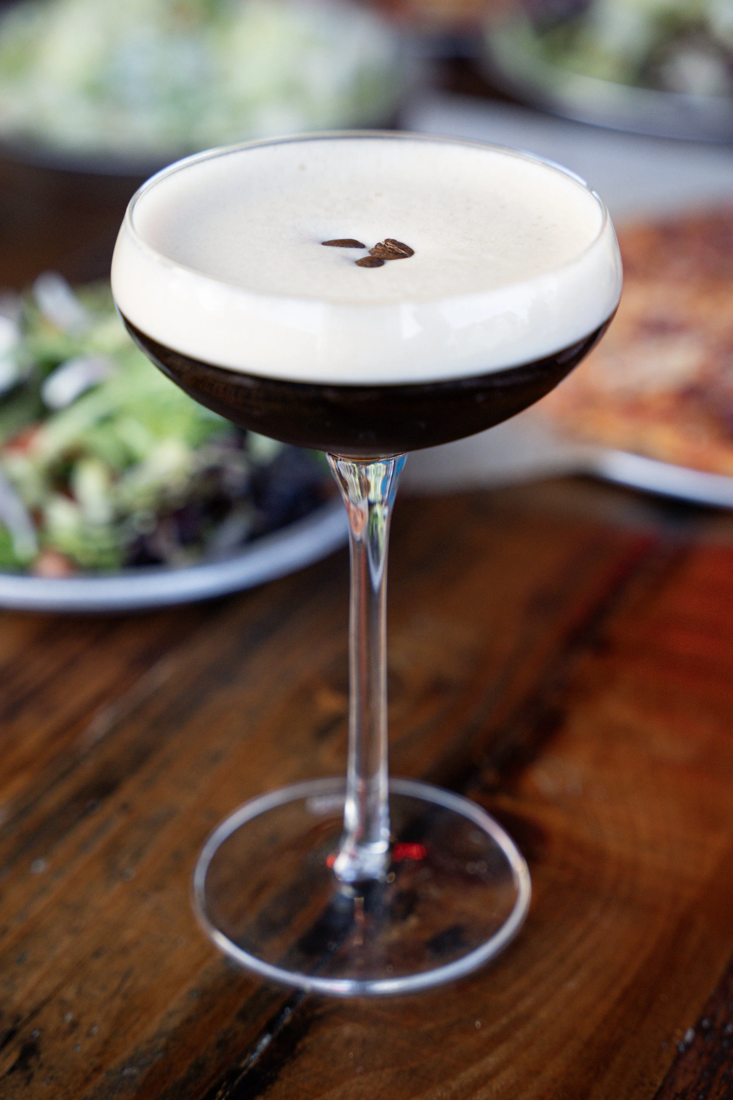 A dark cocktail with a creamy foam top and coffee beans garnish in a coupe glass, placed on a wooden table with a salad in the background.