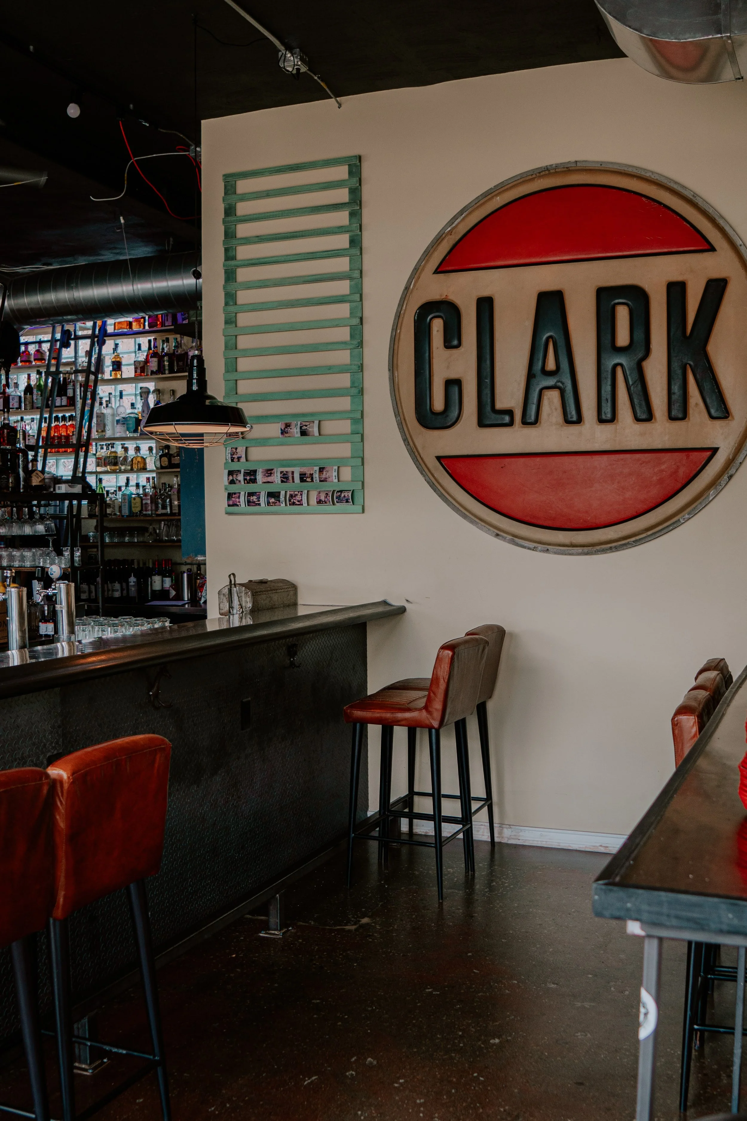 Indoor bar area with a large vintage Clark sign on the wall, high stools with red leather seats, a bar counter, and shelves filled with bottles behind the bar.