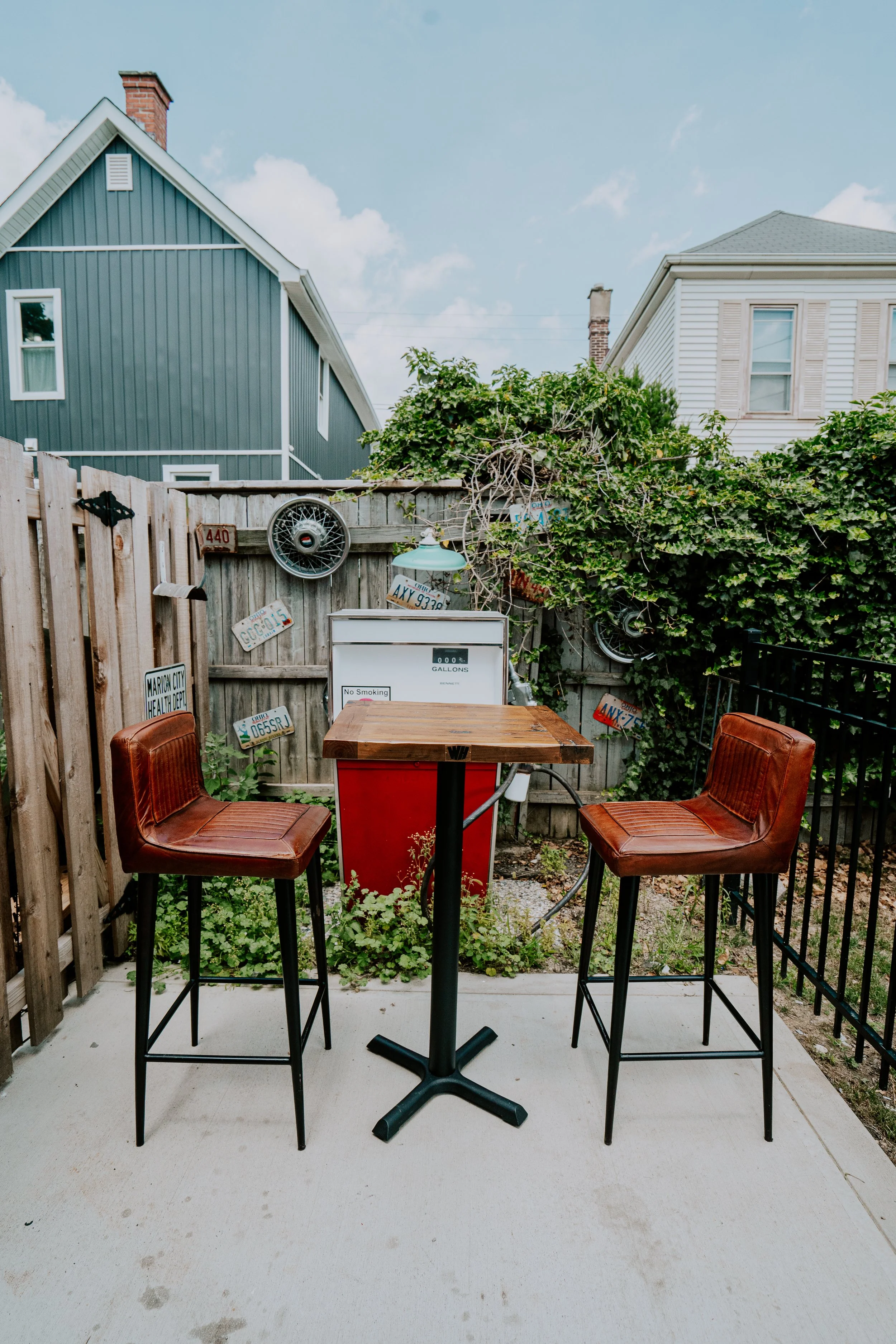 An outdoor patio area with two brown leather bar stools, a high-top table, an old gasoline pump, and a weathered wood fence decorated with vintage license plates. There is greenery and a blue house in the background under a partly cloudy sky.
