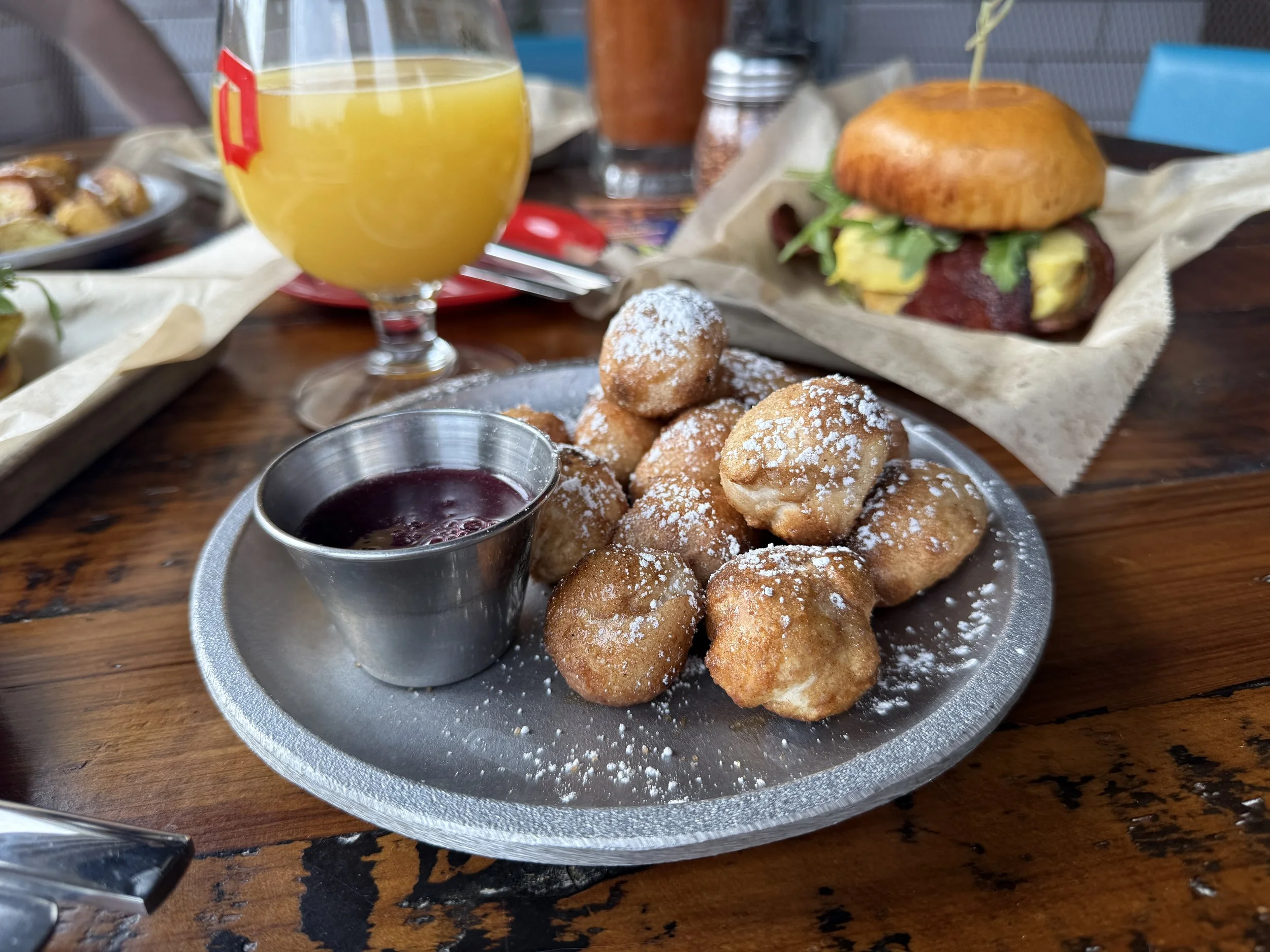 Plate of fried dough balls dusted with powdered sugar and served with berry jam, with a glass of orange juice and a breakfast sandwich in the background.