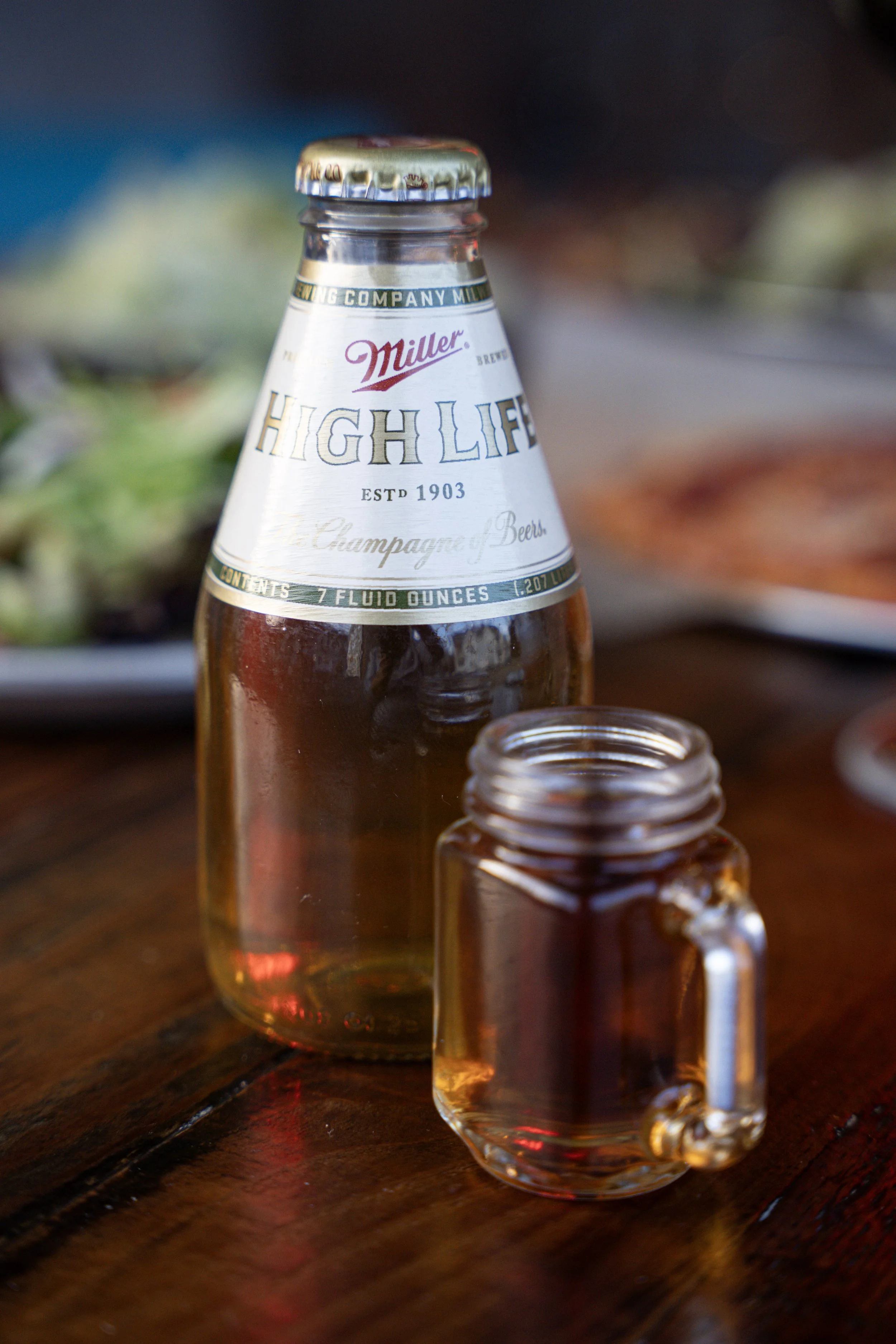 A glass beer bottle labeled 'High Life' and a small glass mug with a handle, both placed on a wooden surface. Blurred dishes are in the background.