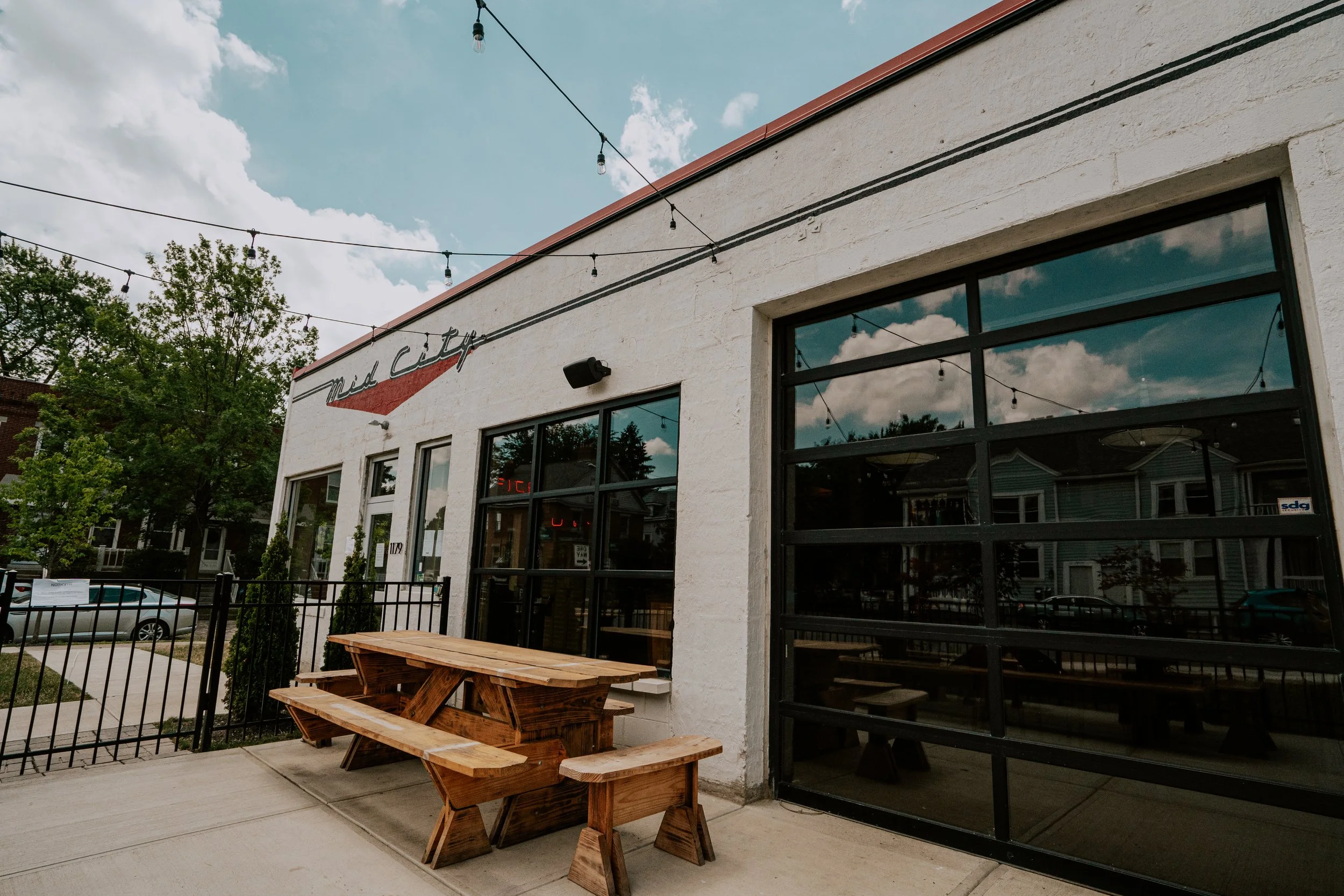 Exterior of a restaurant or cafe called 'Mid City' with large glass windows, outdoor wooden picnic table and benches, string lights overhead, and trees in the background, under a partly cloudy sky.