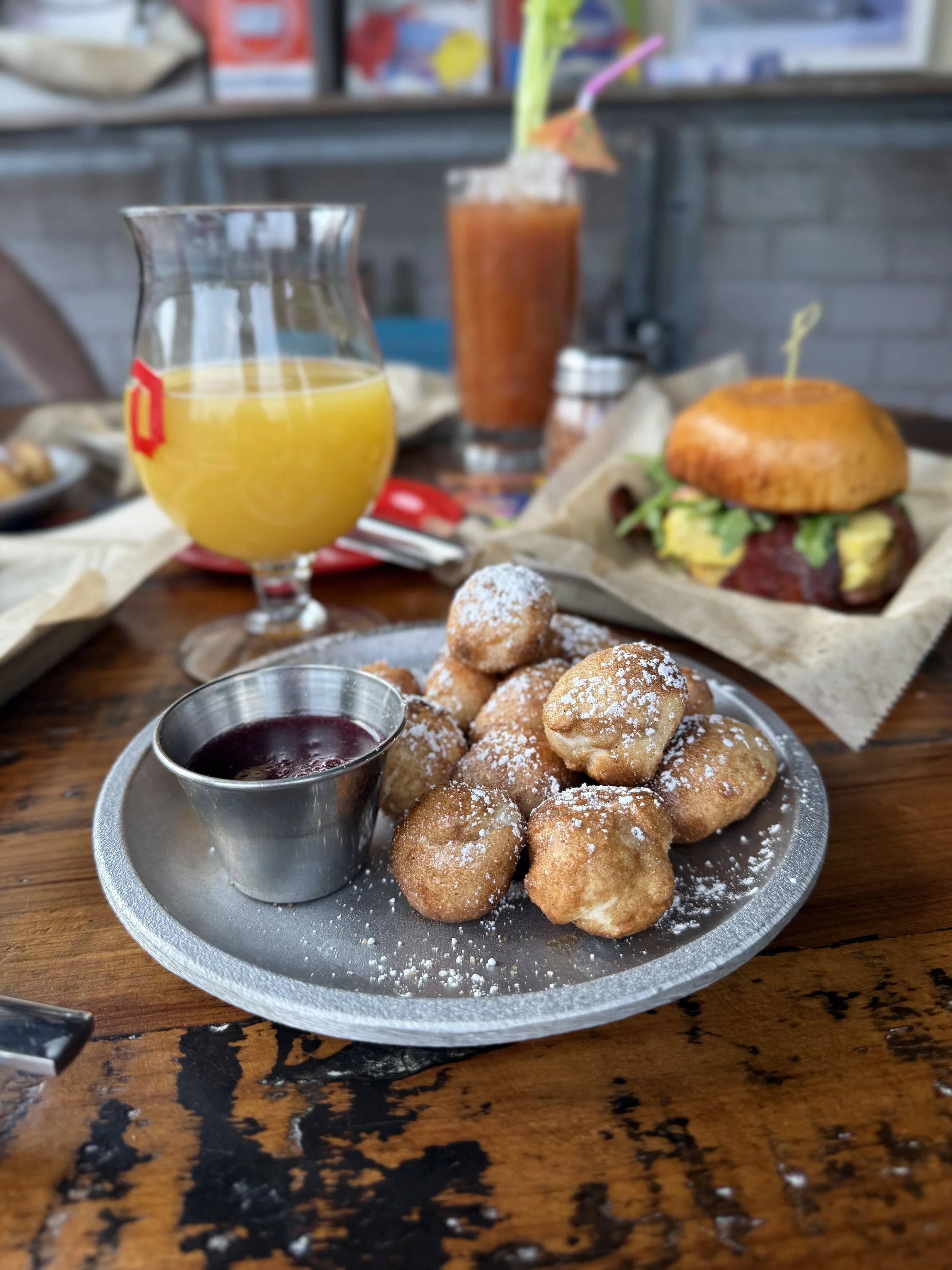 Plate of fried dough balls dusted with powdered sugar and served with a side of berry sauce on a wooden table, with drinks including orange juice, Bloody Mary, and a beer in the background.