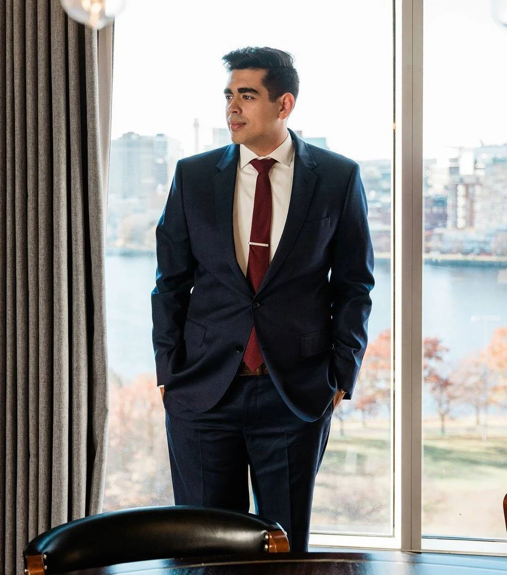 A male attorney in a navy suit with a white shirt and red tie, standing by a large window with a cityscape view.