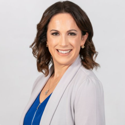 A woman with shoulder-length dark brown hair, wearing a light gray blazer over a blue top, smiling against a plain light gray background.