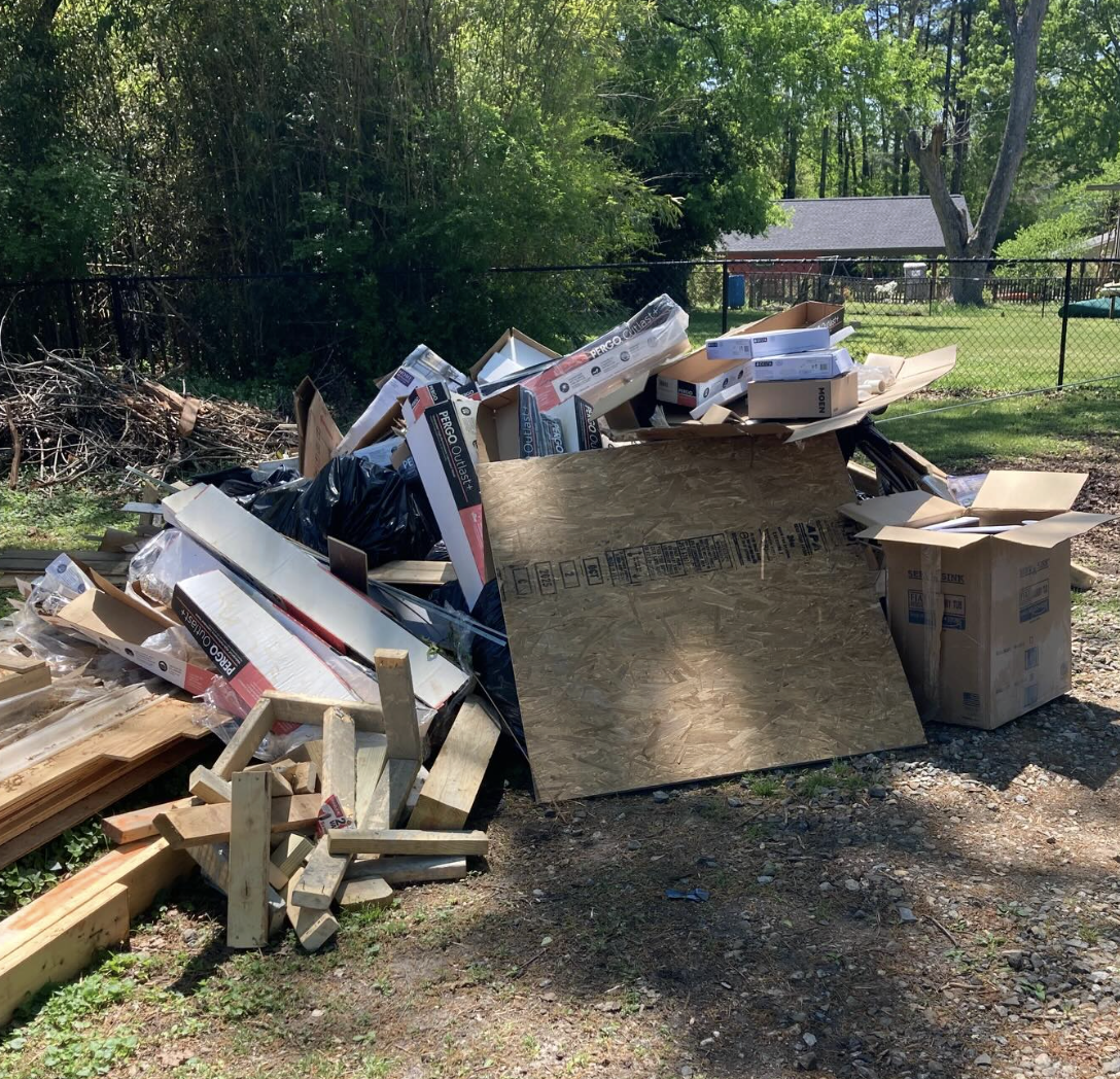 Pile of discarded cardboard boxes, packed wood, and trash outdoors on the ground near a chain-link fence and trees.