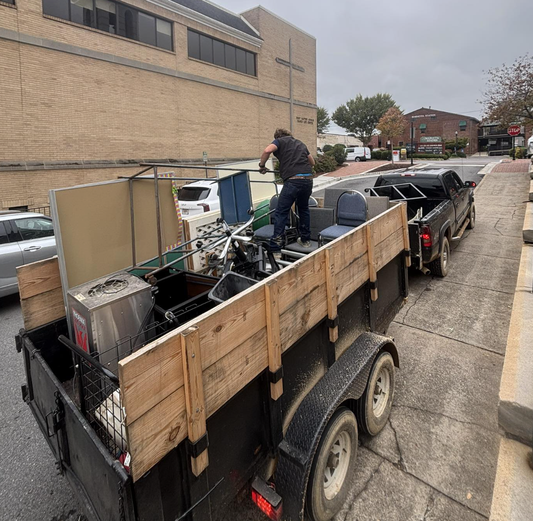 A man loading items onto a trailer attached to a black pickup truck parked on a city street overcast day, with a church with a cross on the wall visible in the background.