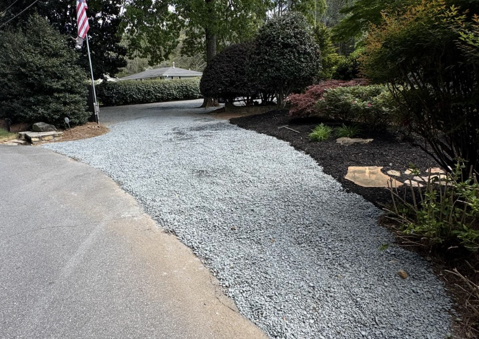 A gravel driveway surrounded by landscaped bushes and trees, with a small American flag on a pole in the background.