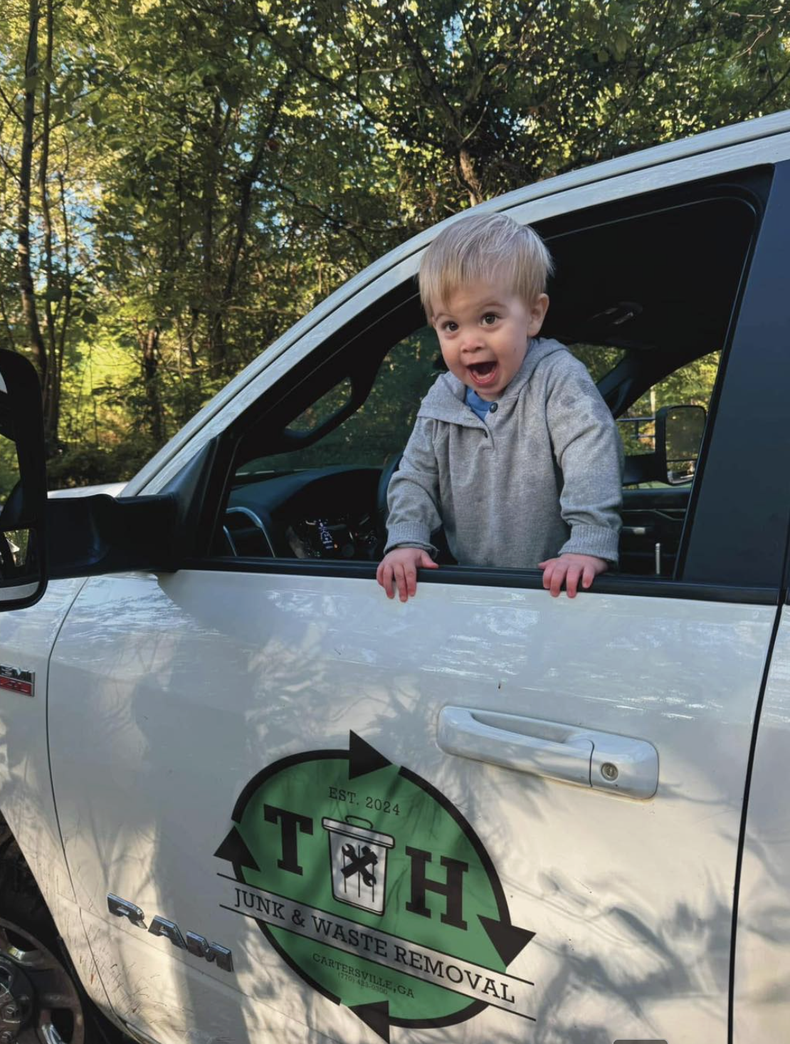 A young boy with blonde hair leaning out of a white truck window, smiling with his mouth open, surrounded by green trees.