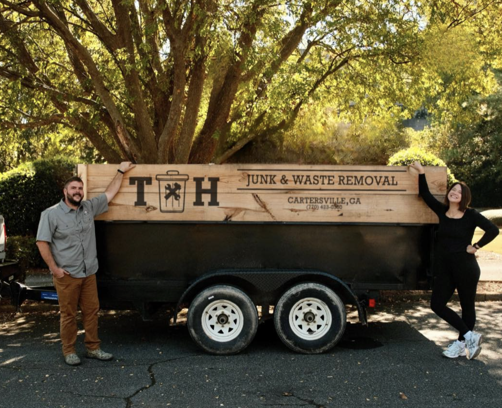 Two people stand on either side of a trailer with a large wooden sign for a junk and waste removal business, with a large tree in the background.