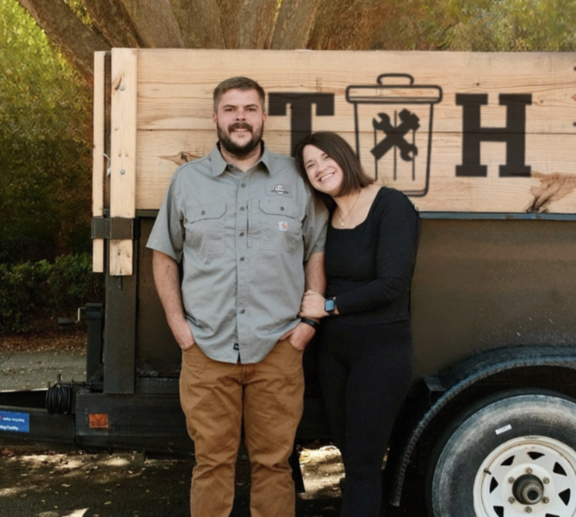 A man and woman stand close together, smiling and posing in front of a wooden sign with a trash can graphic and large black letters, outdoors with trees in the background.