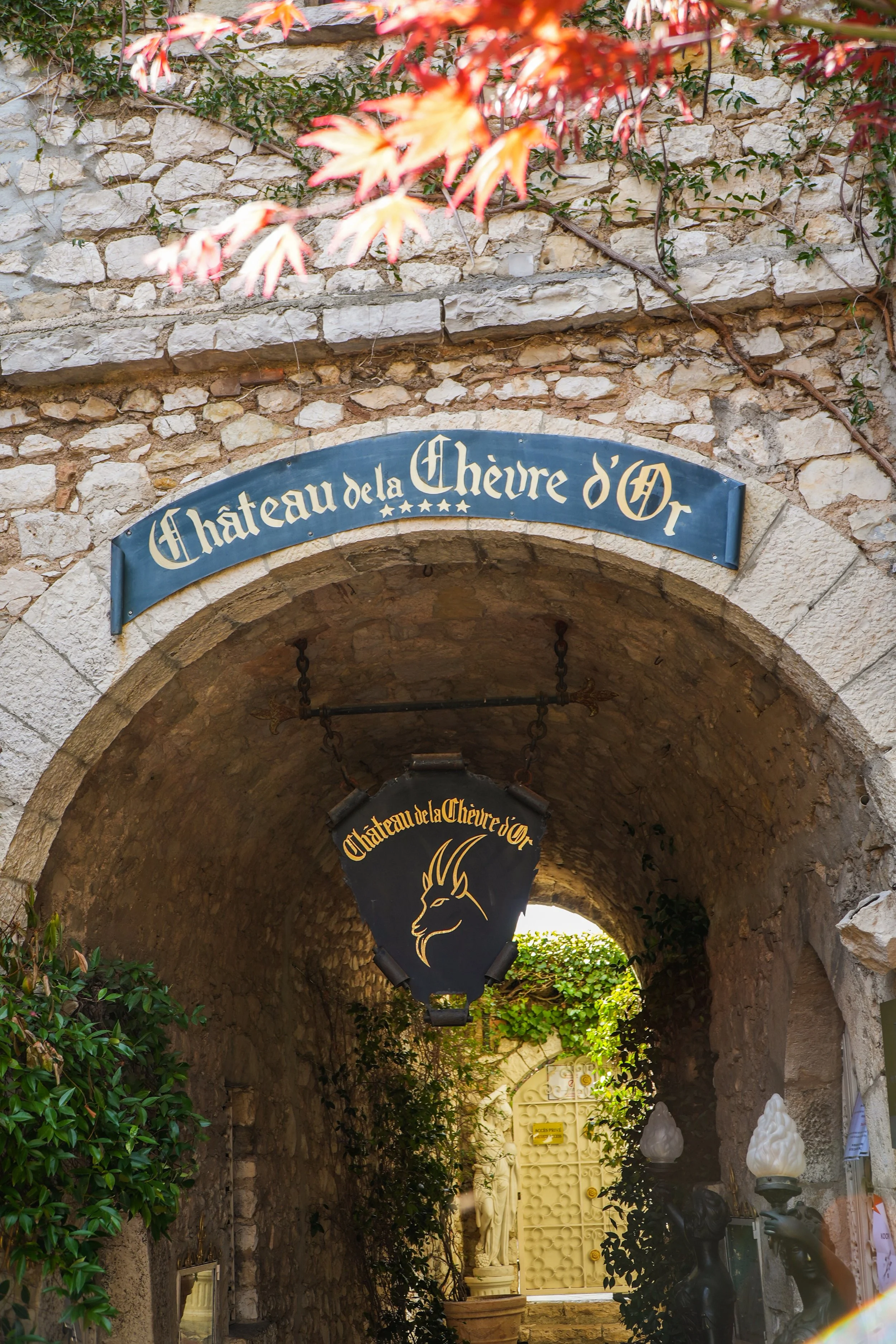 Entrée d'un château en pierre avec une arche et une enseigne en forme de bouclier noir portant un dessin d'un chèvre et le texte 'Château de la Chèvre d'Or', entourée de végétation et statues de dragons.