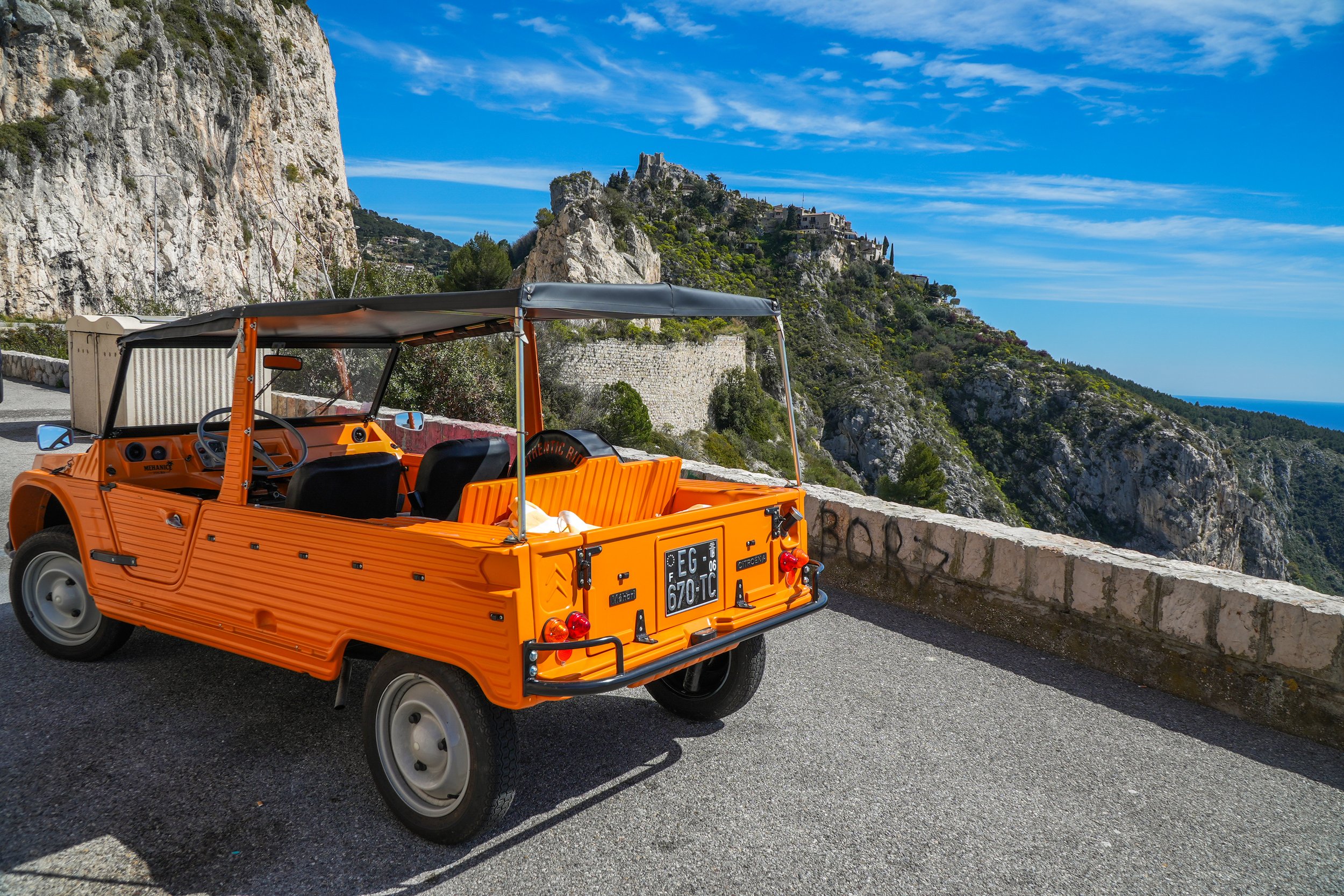 Voiture orange vintage avec un toit en toile, stationnée le long d'une route avec un paysage de collines rocheuses et de maisons en arrière-plan, sous un ciel bleu avec quelques nuages.