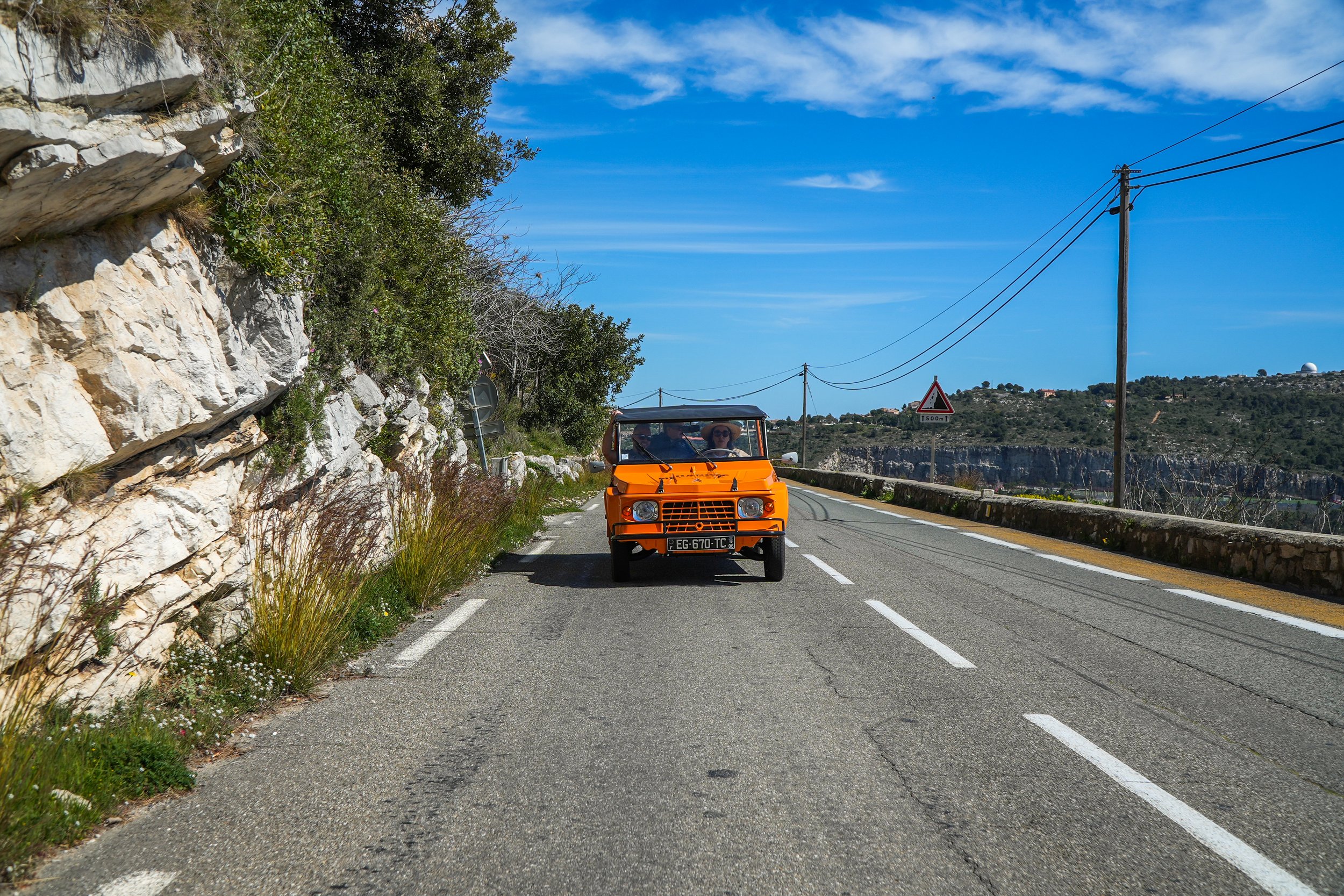 Voiture orange ancienne sur une route de campagne avec un ciel bleu et des collines en arrière-plan.