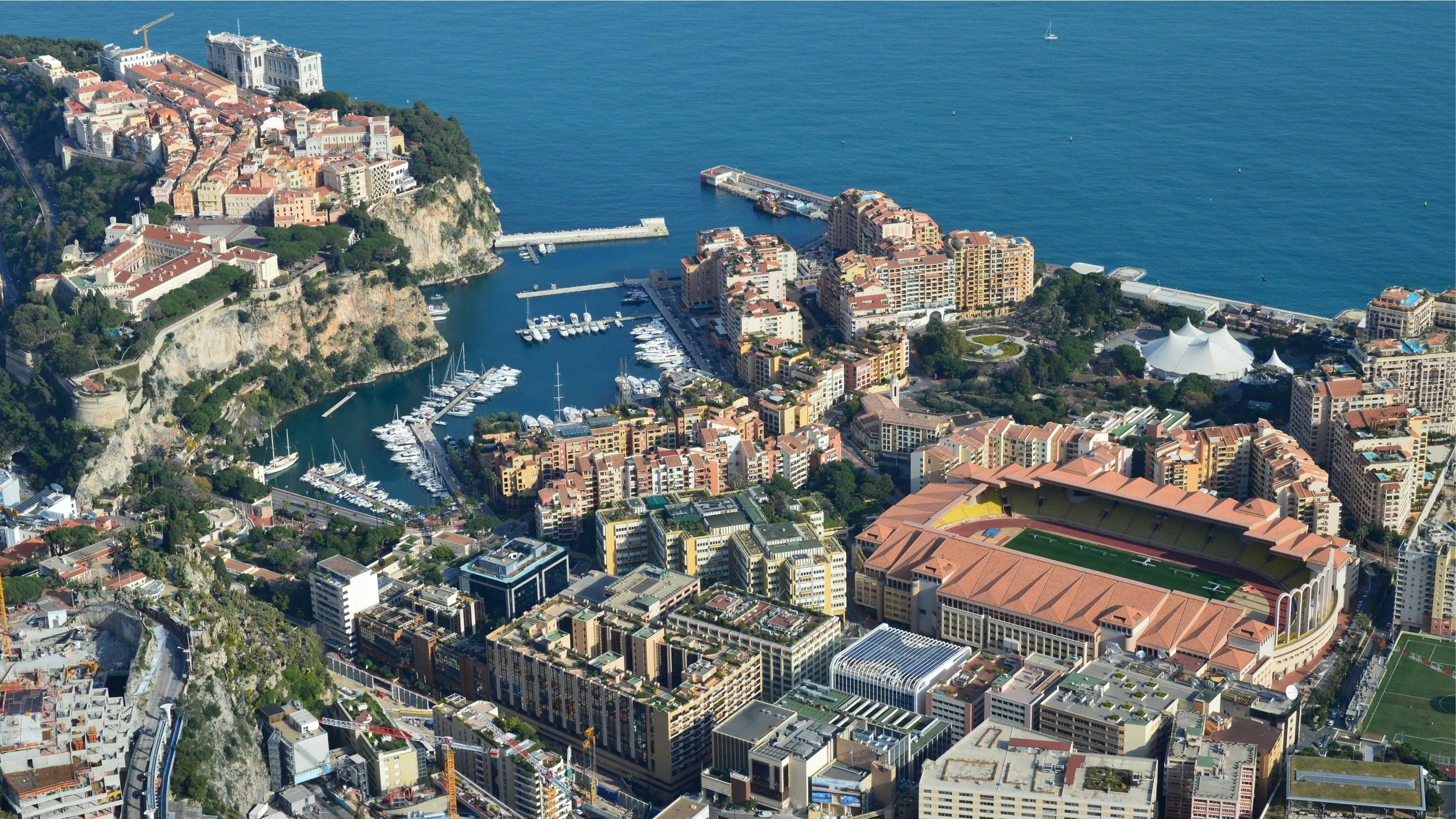 Vue aérienne de Monaco avec son port, ses bâtiments résidentiels et commerciaux, le stade de football, le palais princier en haut, et la Méditerranée en arrière-plan.