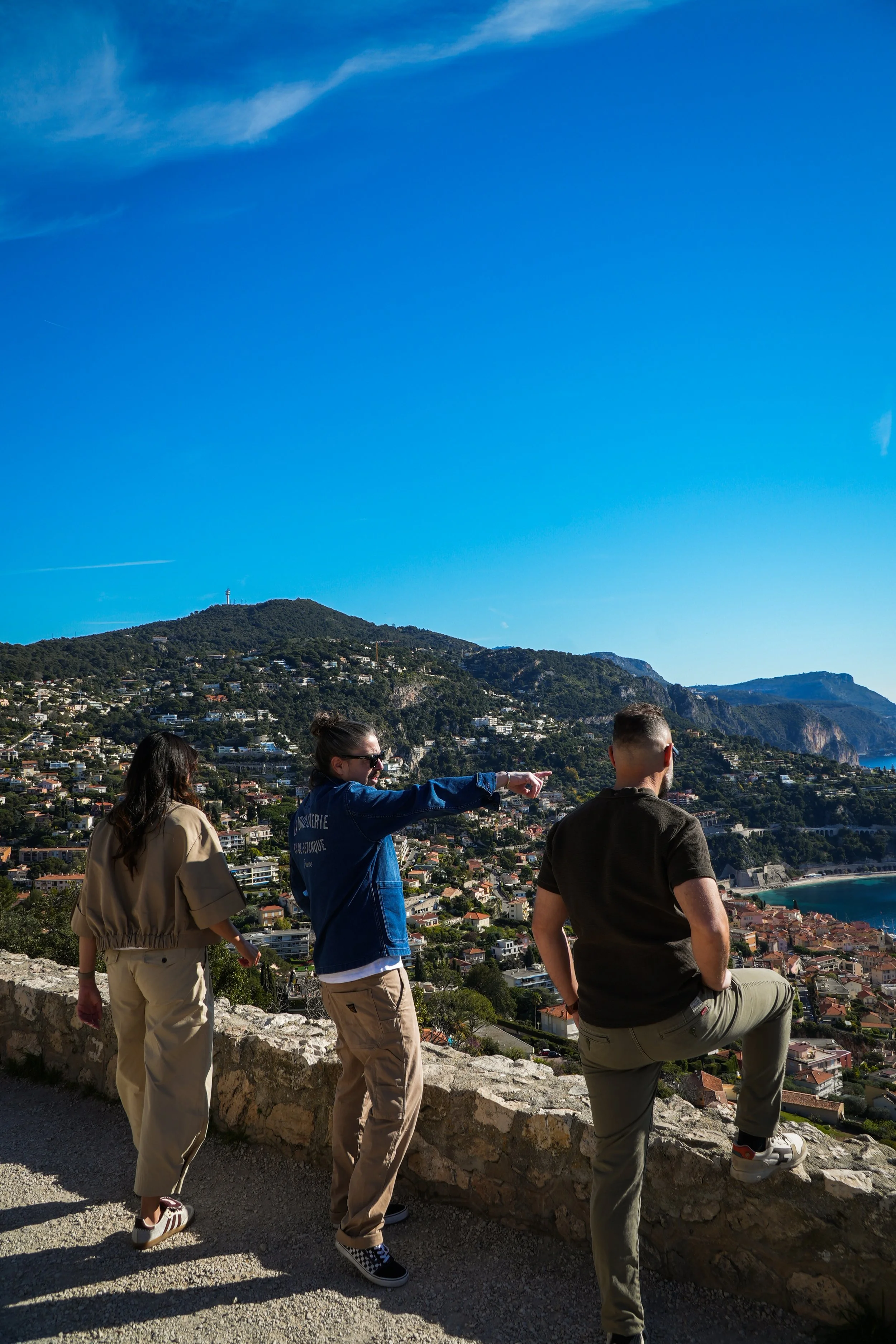 Quatre personnes âgées d'environ 20 à 30 ans sont debout sur un chemin en hauteur, face à une ville côtière et une mer. L'une d'elles, une femme, pointe du doigt quelque chose dans la ville. Le paysage est ensoleillé, avec une montagne en arrière-plan sous un ciel bleu clair.