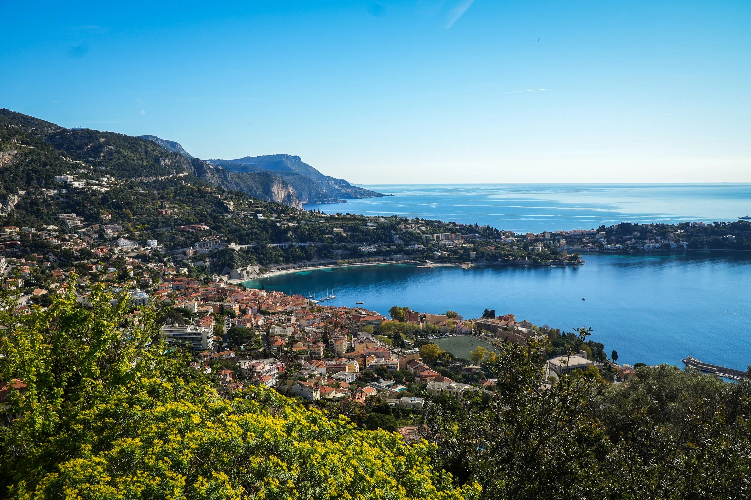 Vue panoramique sur la côte méditerranéenne avec une ville en contrebas, des montagnes boisées à gauche et une mer bleue à droite, sous un ciel clair.