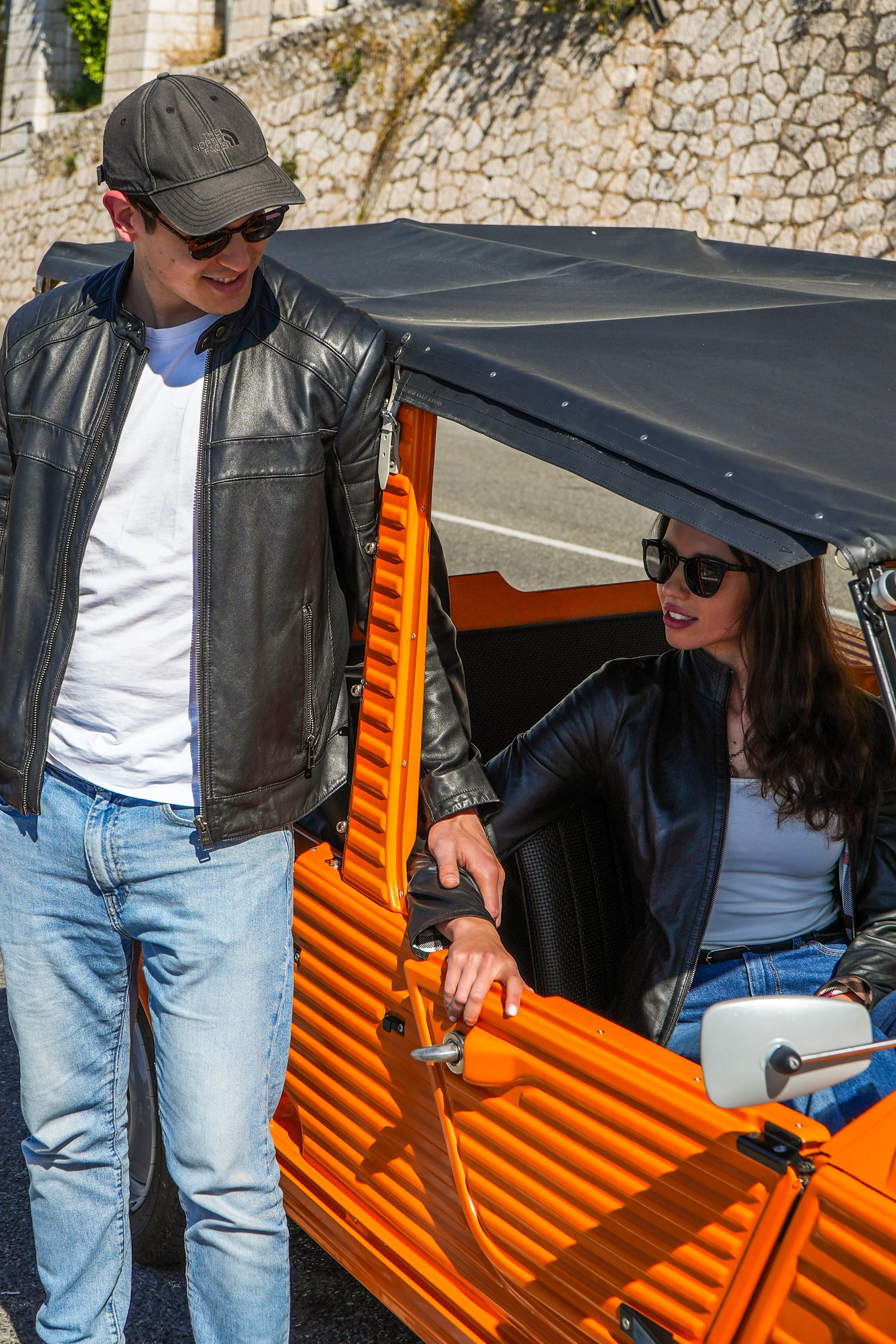 Un homme et une femme portant des lunettes de soleil et des vêtements en cuir, debout près d'un véhicule à triporteur orange, sur fond de mur en pierre.