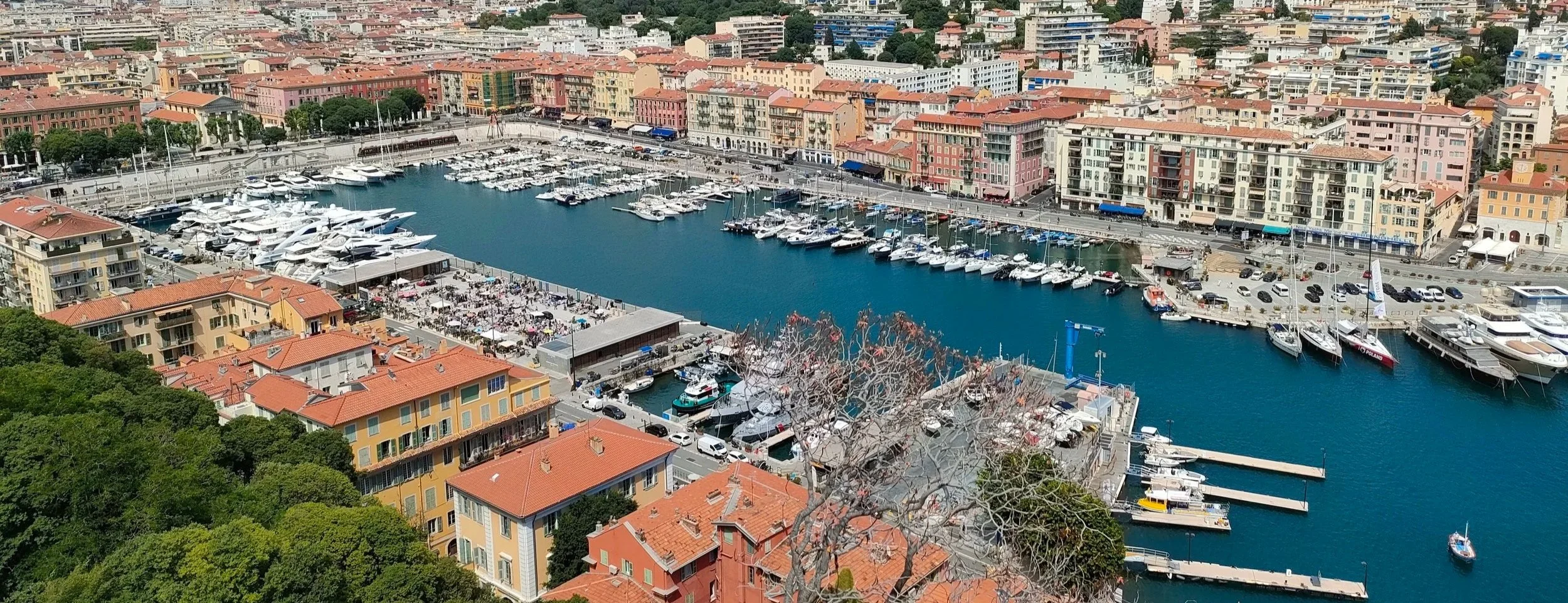 Port marina avec de nombreux yachts et voiliers, entouré de bâtiments résidentiels colorés, dans une ville en pente avec des rues animées et de la verdure