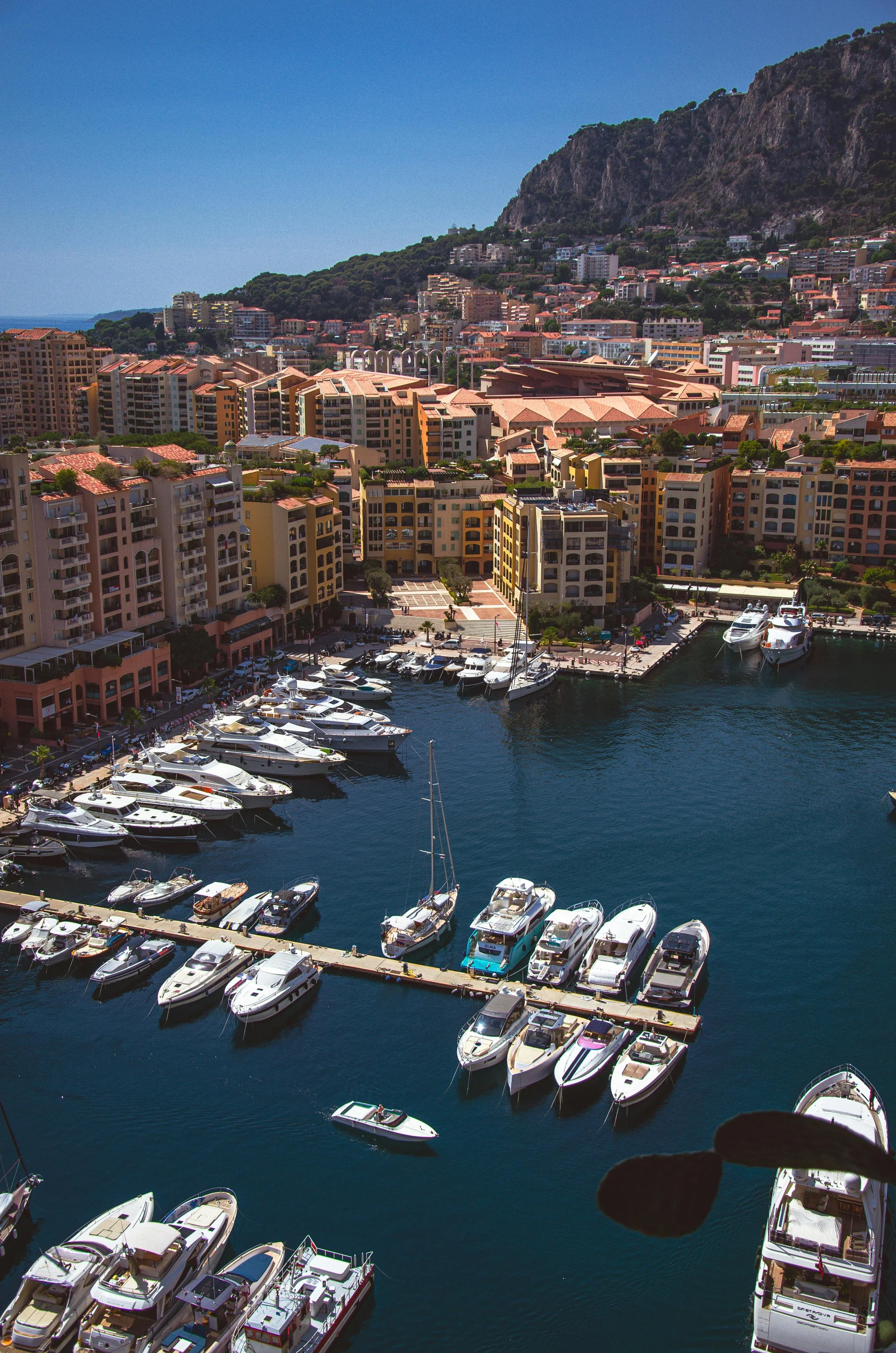 Port de yachts avec de nombreux bateaux amarrés, bâtiments résidentiels et montagnes en arrière-plan sous ciel clair.