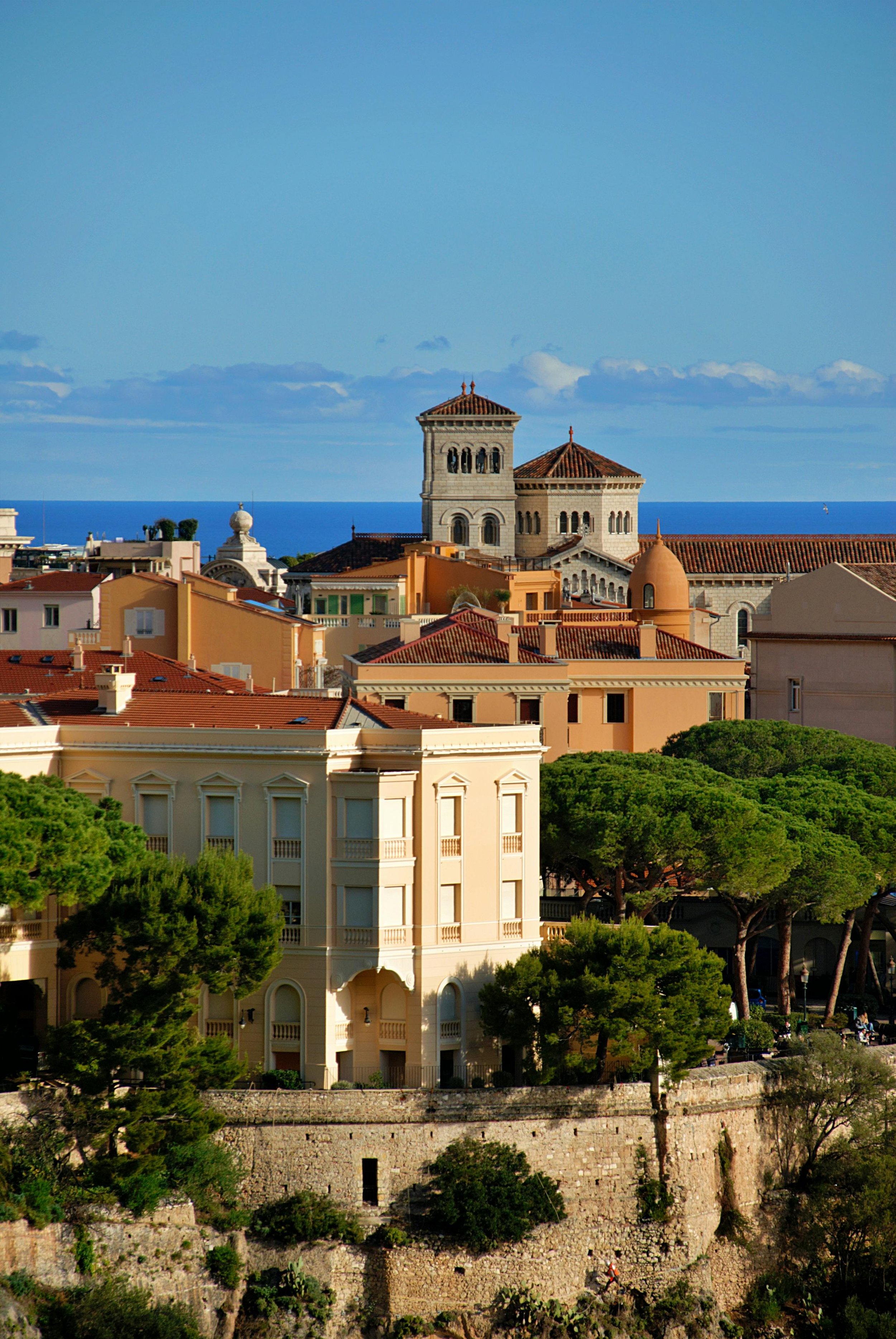 Vue d'une ville côtière avec des bâtiments de style méditerranéen, un clocher d'église en pierre, et la mer en arrière-plan sous un ciel bleu avec quelques nuages.