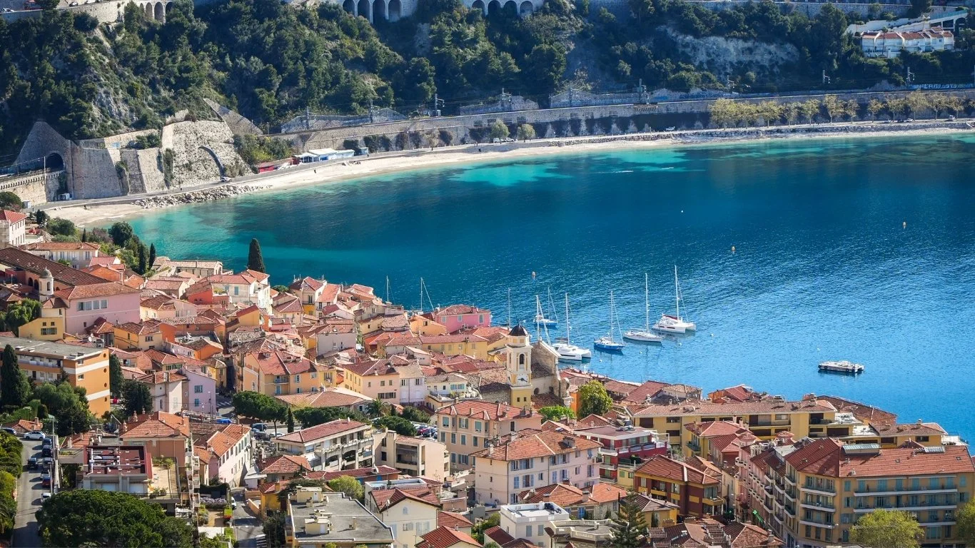 Cliché d'une ville côtière avec des maisons colorées, des yachts au quai et une mer turquoise entourée de montagnes vertes.