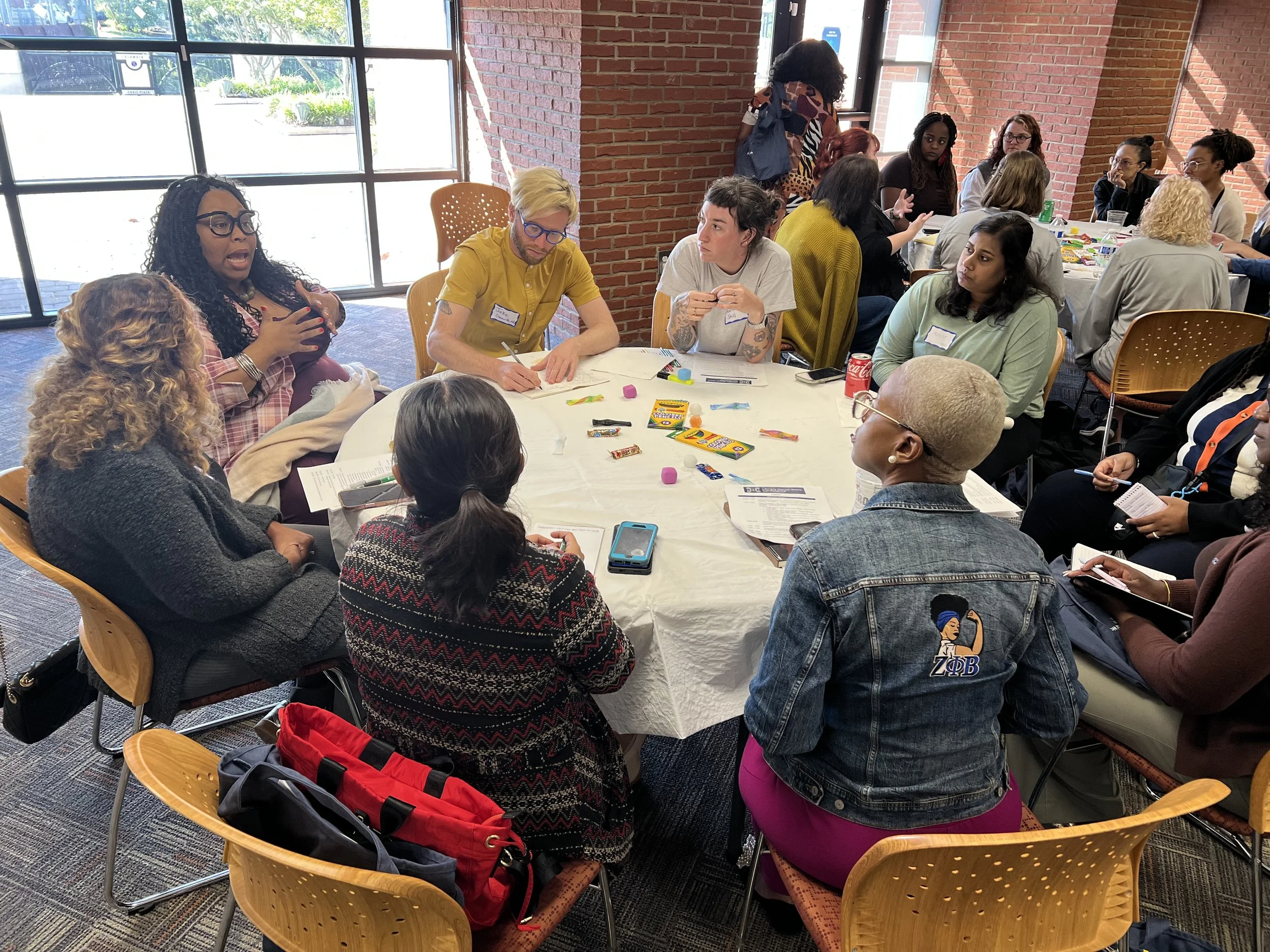 A group of diverse women and men sitting around a round table engaged in discussion during a workshop or meeting in a bright room with large windows and brick walls.