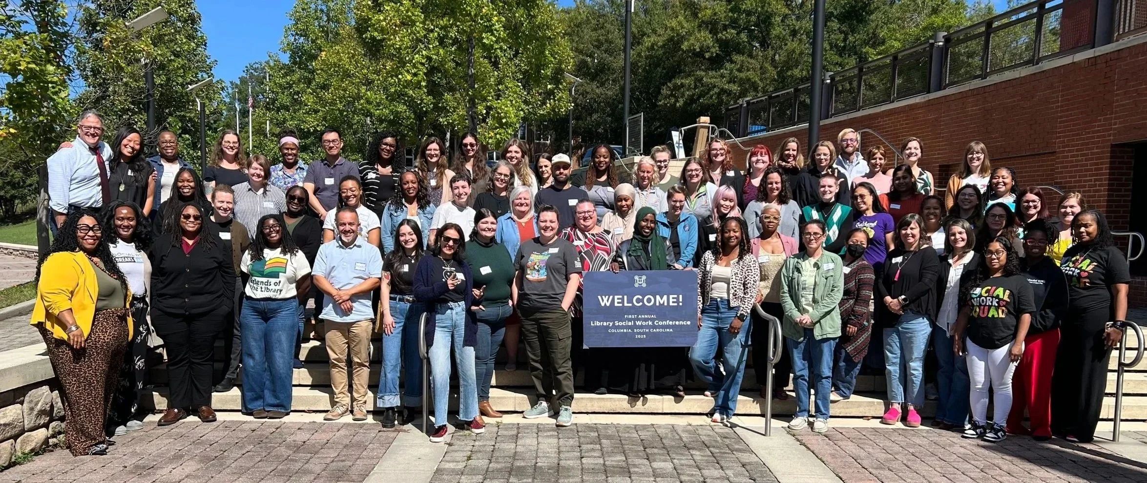 A large group of diverse people gathered outdoors for a photo at the Library Social Work Conference in Columbia, South Carolina, 2023. They are standing on steps behind a welcome sign, with trees and a brick building in the background.