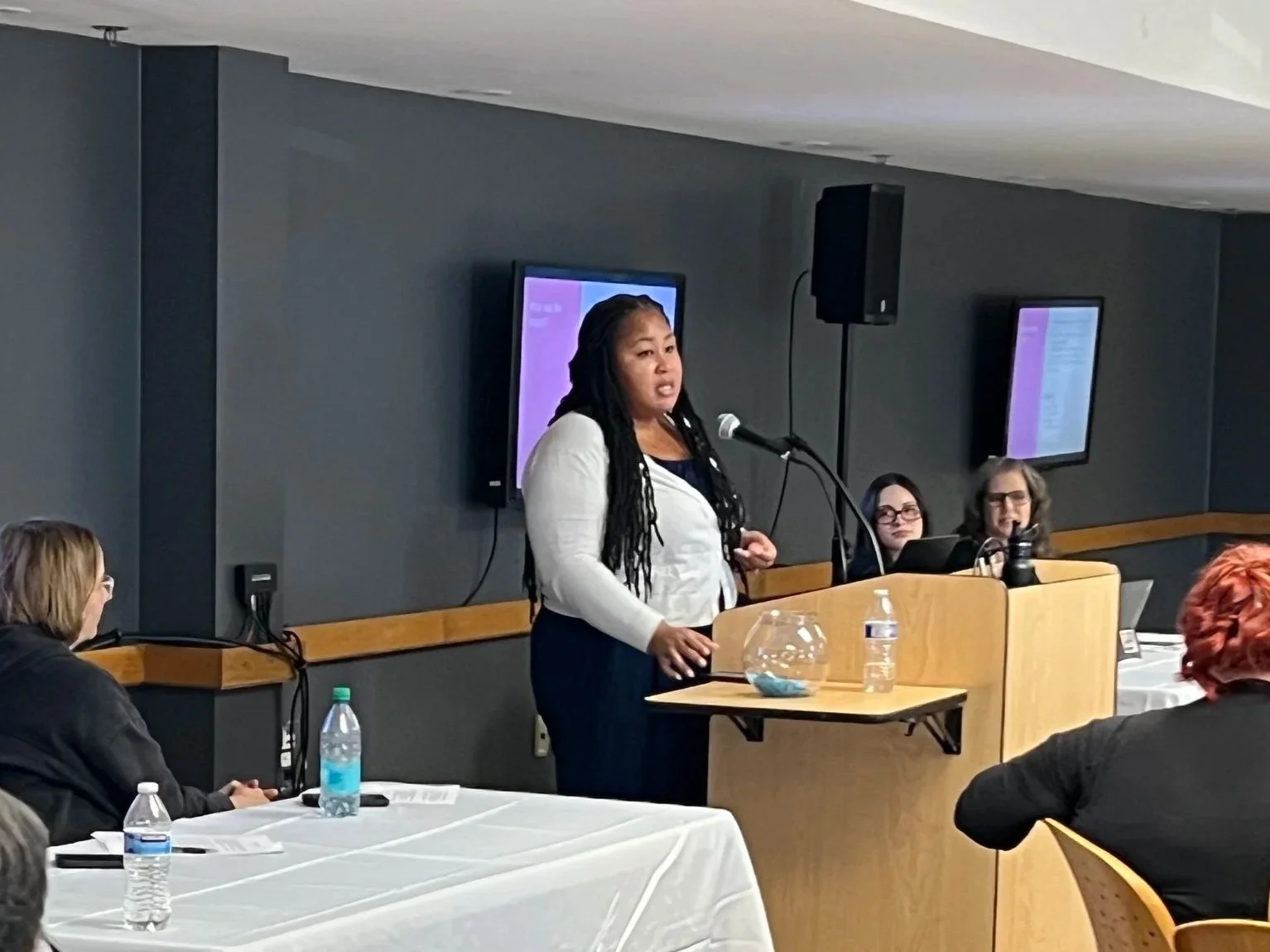 A woman with long braids speaking at a podium during a presentation or conference, with three women seated behind her and two people visible in the foreground, in a room with dark walls and two screens on the wall.