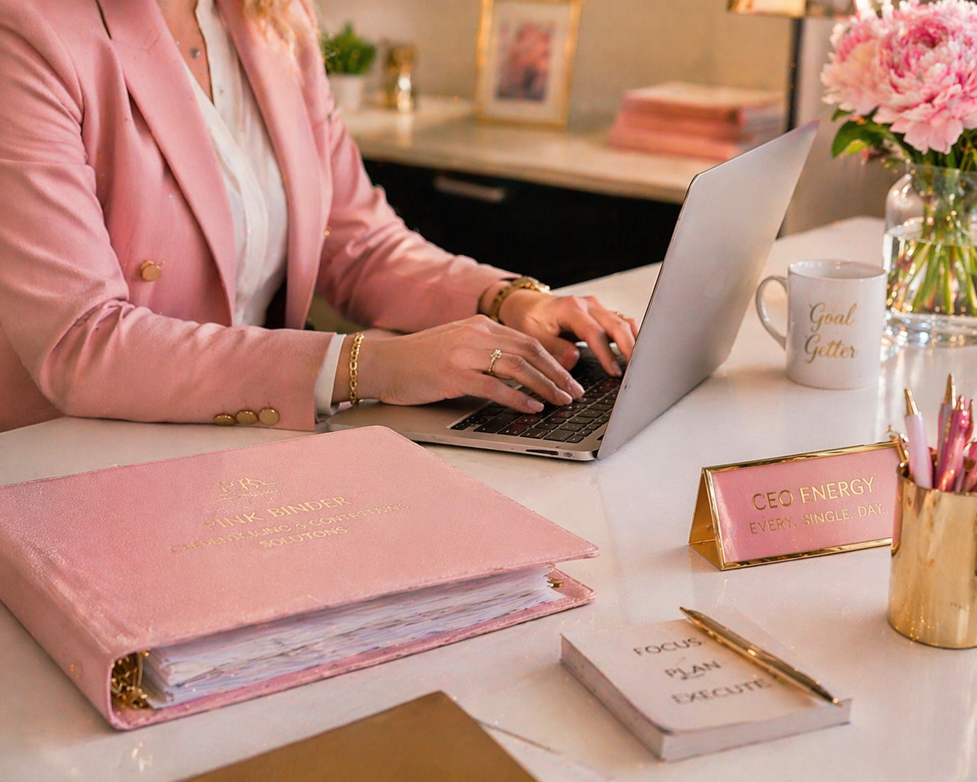 A woman in a pink blazer working on a laptop at a white desk. The desk has a pink binder labeled 'Pink Binder, Resume & Covering Solutions', a nameplate reading 'CEO Energy, Every. Single. Day.', a white mug with 'Goal Getter' written on it, a potted plant with pink flowers, a pen holder with pink pens, and a notebook. The background shows framed pictures and a stack of pink books.