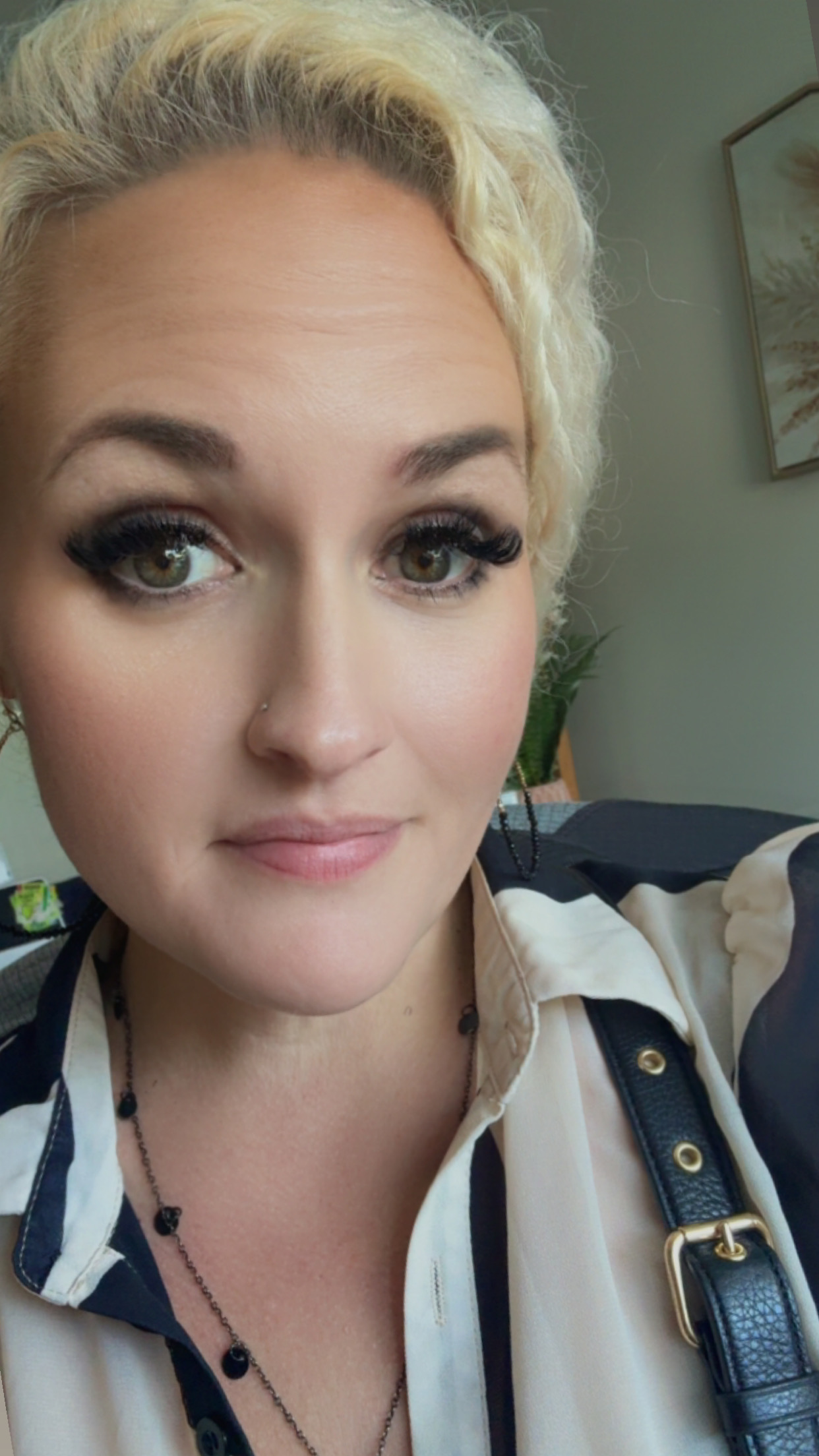 Close-up of a woman with blonde curly hair, gray eyes, makeup, a small nose stud, wearing earrings, a necklace, a cream and black shirt, and a black strap with a gold buckle.