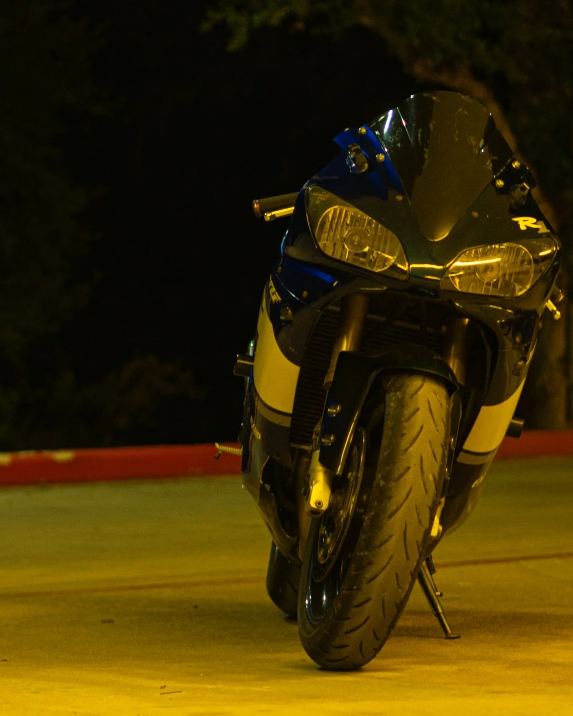 A sport motorcycle parked on a concrete surface at night, facing slightly downward, with a dark background and trees in the distance.
