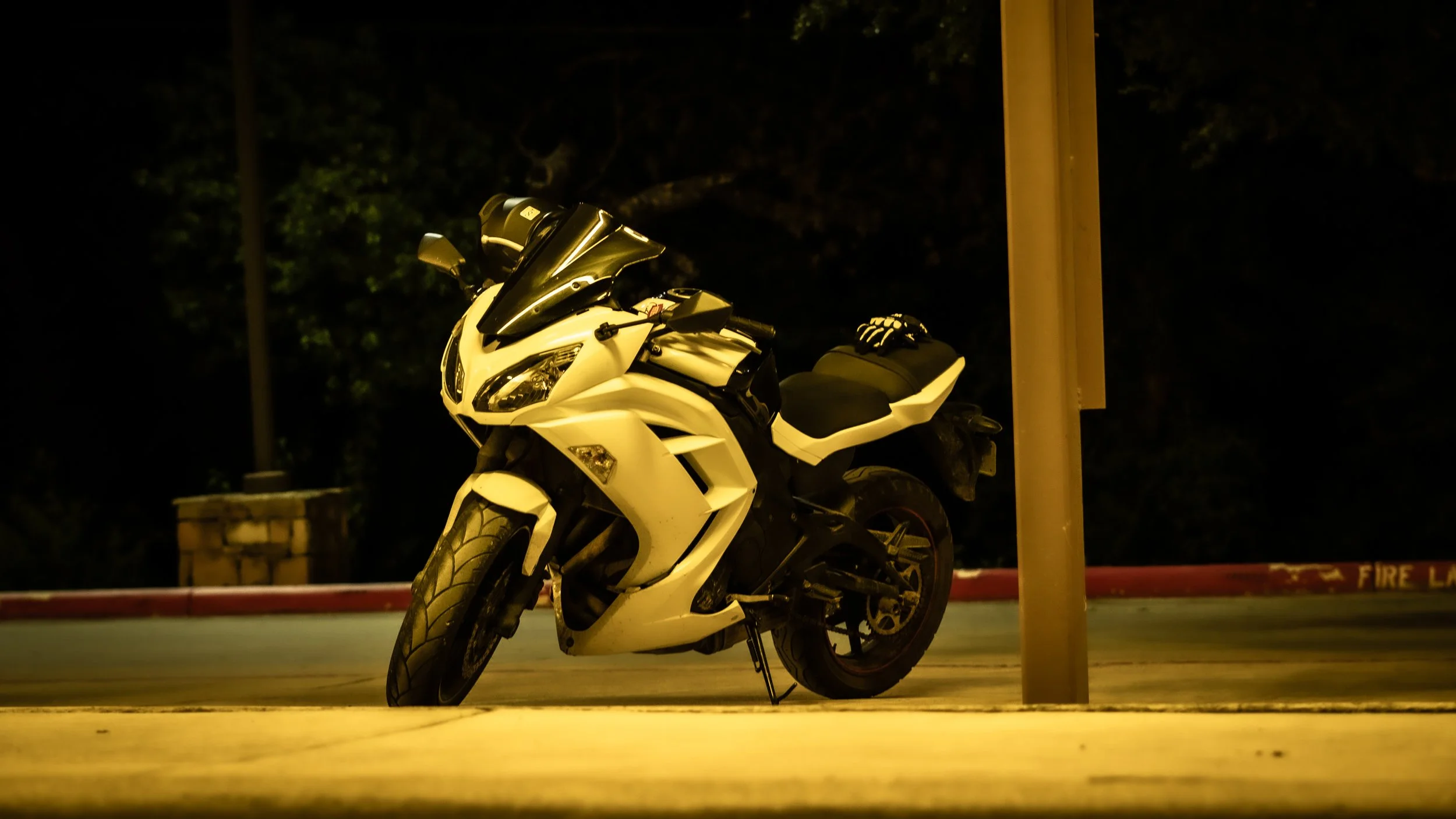A white sports motorcycle parked on a street at night, with a black helmet on the seat and a pair of gloves nearby, illuminated by streetlights.