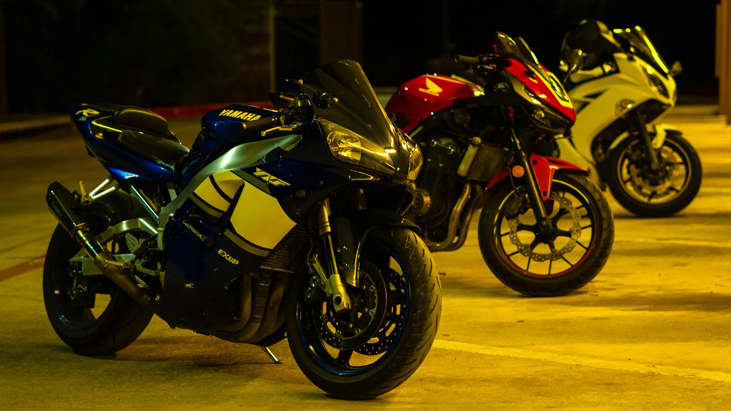 Three sport motorcycles parked in a dimly lit parking lot at night, with the closest one painted in black, blue, and yellow, followed by red and white, and yellow and black bikes.