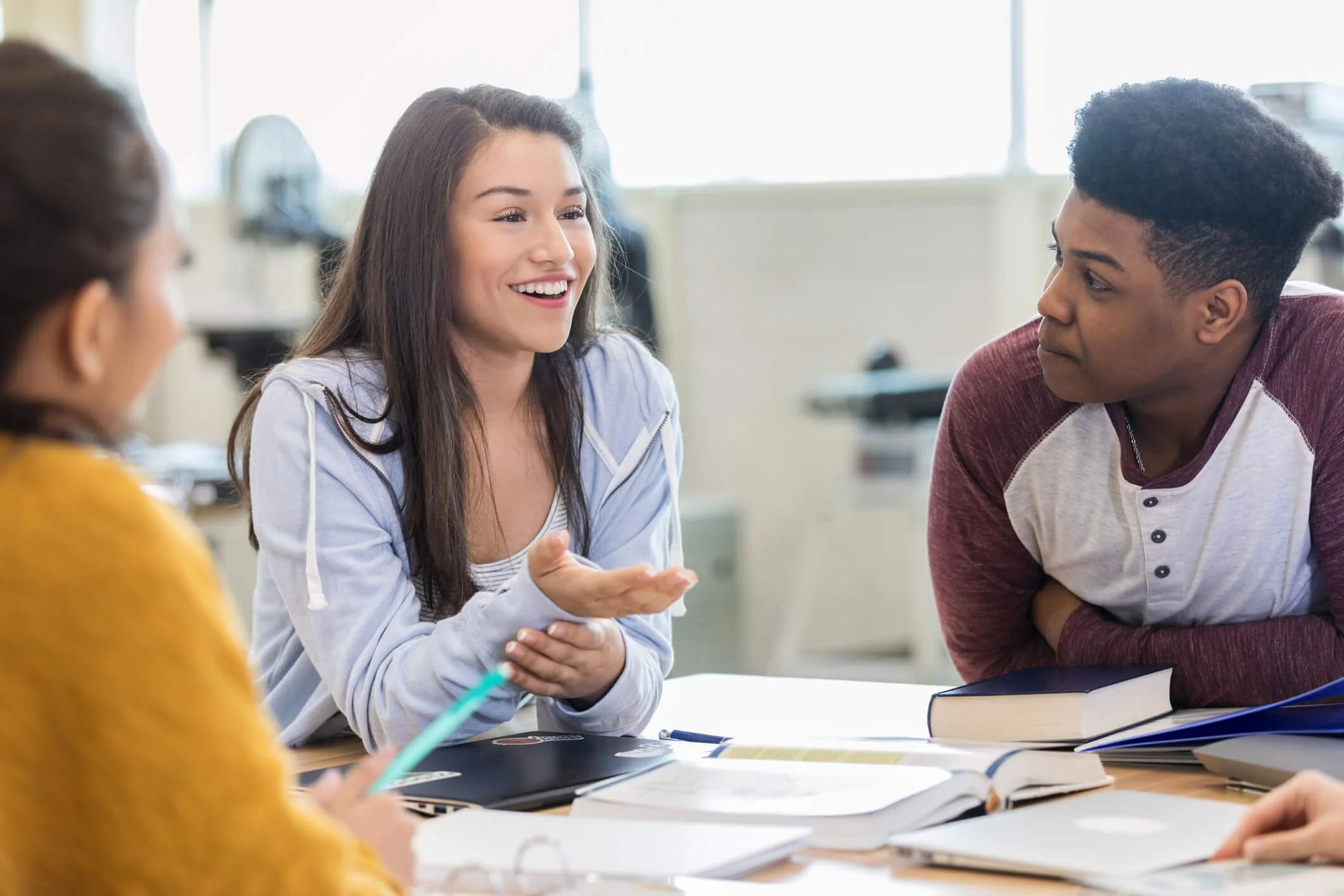 Three students engaged in a lively conversation in a classroom setting, with notebooks and textbooks on the table.
