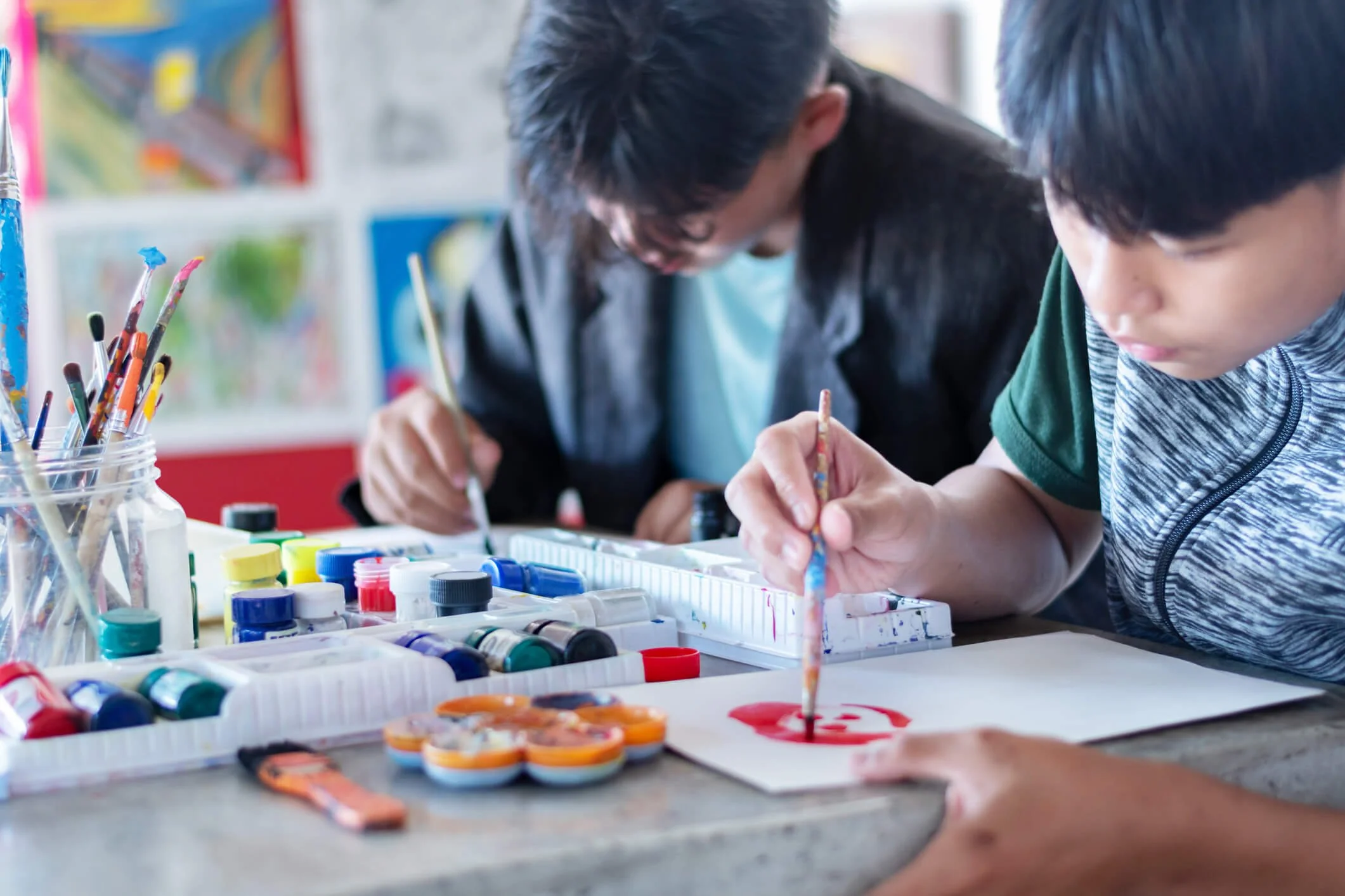 Two children painting with watercolors at a table, surrounded by various paint supplies.