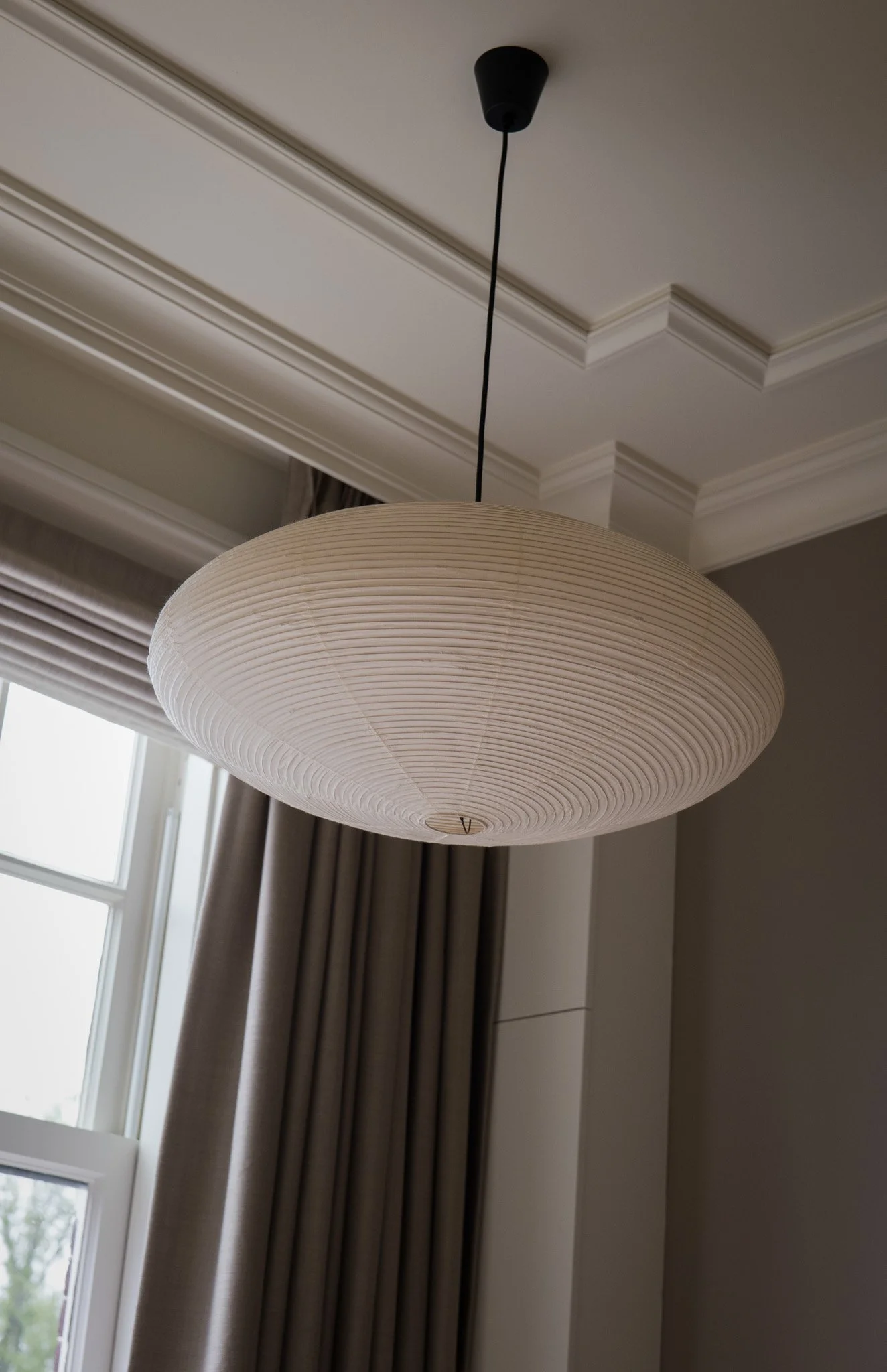 Witte paper-lamp hangend aan het plafond in een woonkamer met grote ramen bedekt met beige en grijze gordijnen.