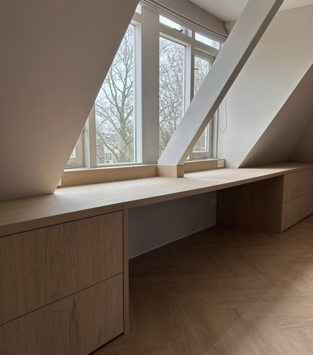 Lijnsgevel met houten bureau onder schuine ramen tot het plafond, binnen in een kamer met houten vloer en zicht op een boom buiten.