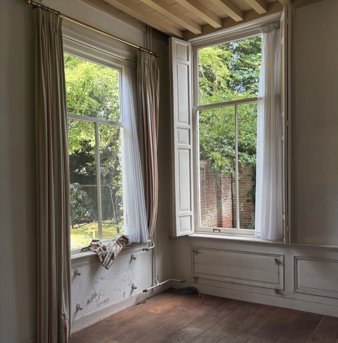 Een kamer met twee grote ramen, een met gordijnen en een zonder. Buiten de ramen is groen landschap en een muur zichtbaar.