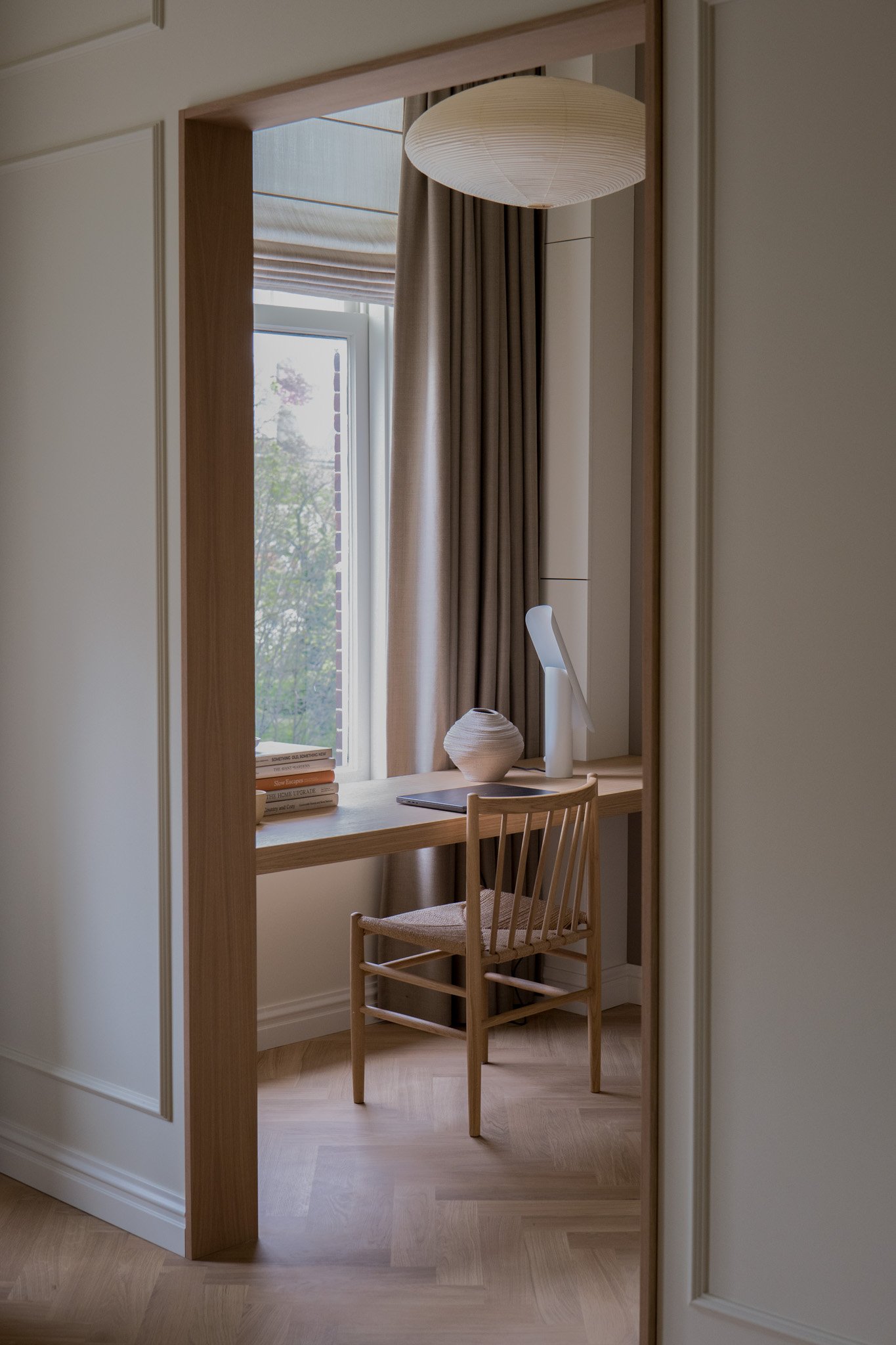 Een knusse werkhoek met een houten bureau, een houten stoel, een stapel boeken, een vaas, een bureau- of tafellamp, een raam met beige gordijnen en een grote papieren hanglamp.