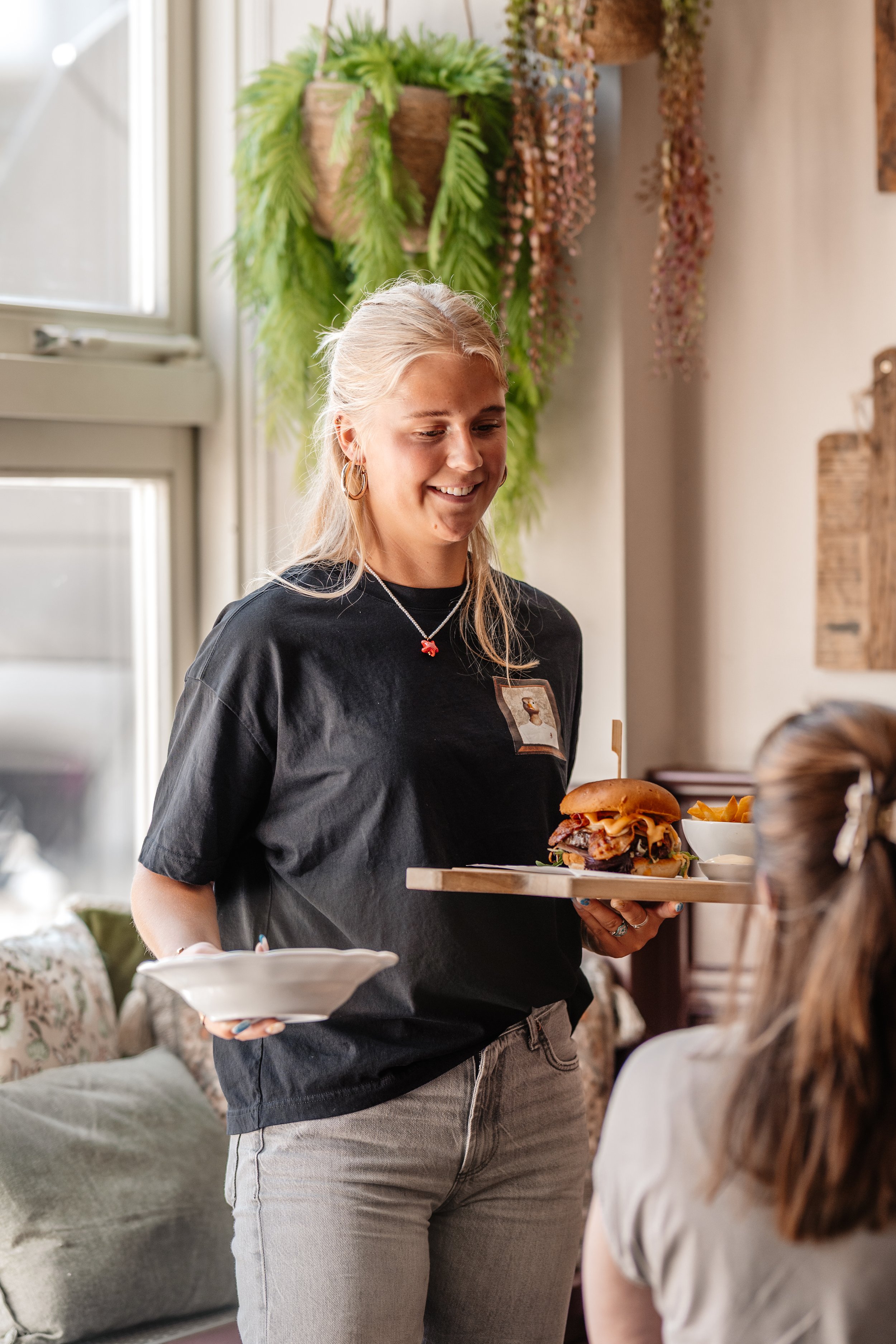 A woman with blonde hair wearing a black t-shirt serving a burger on a platter to someone seated at a table in a cozy, decorated restaurant.