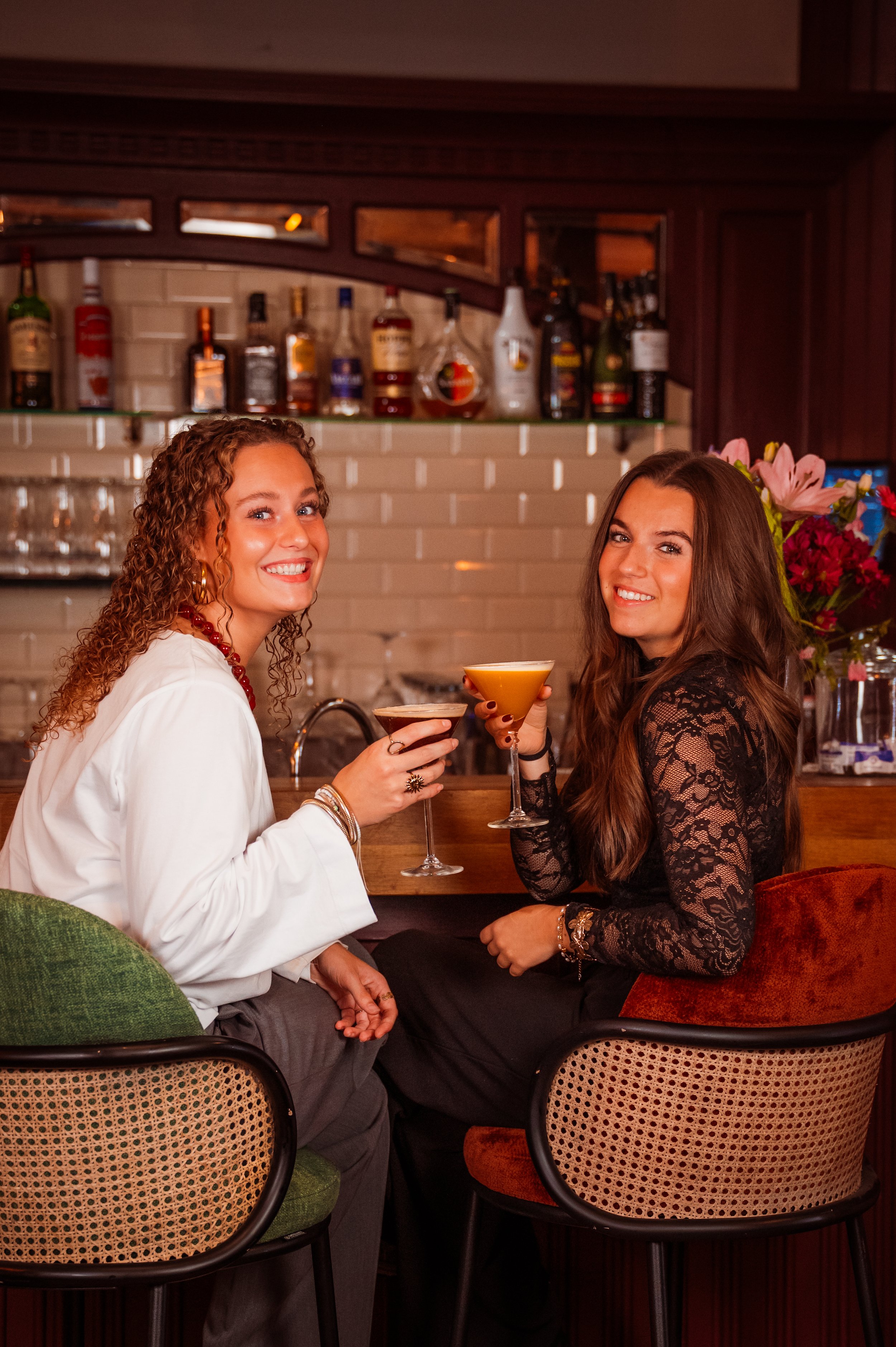 Two women sitting at a bar, holding cocktails, smiling at the camera.