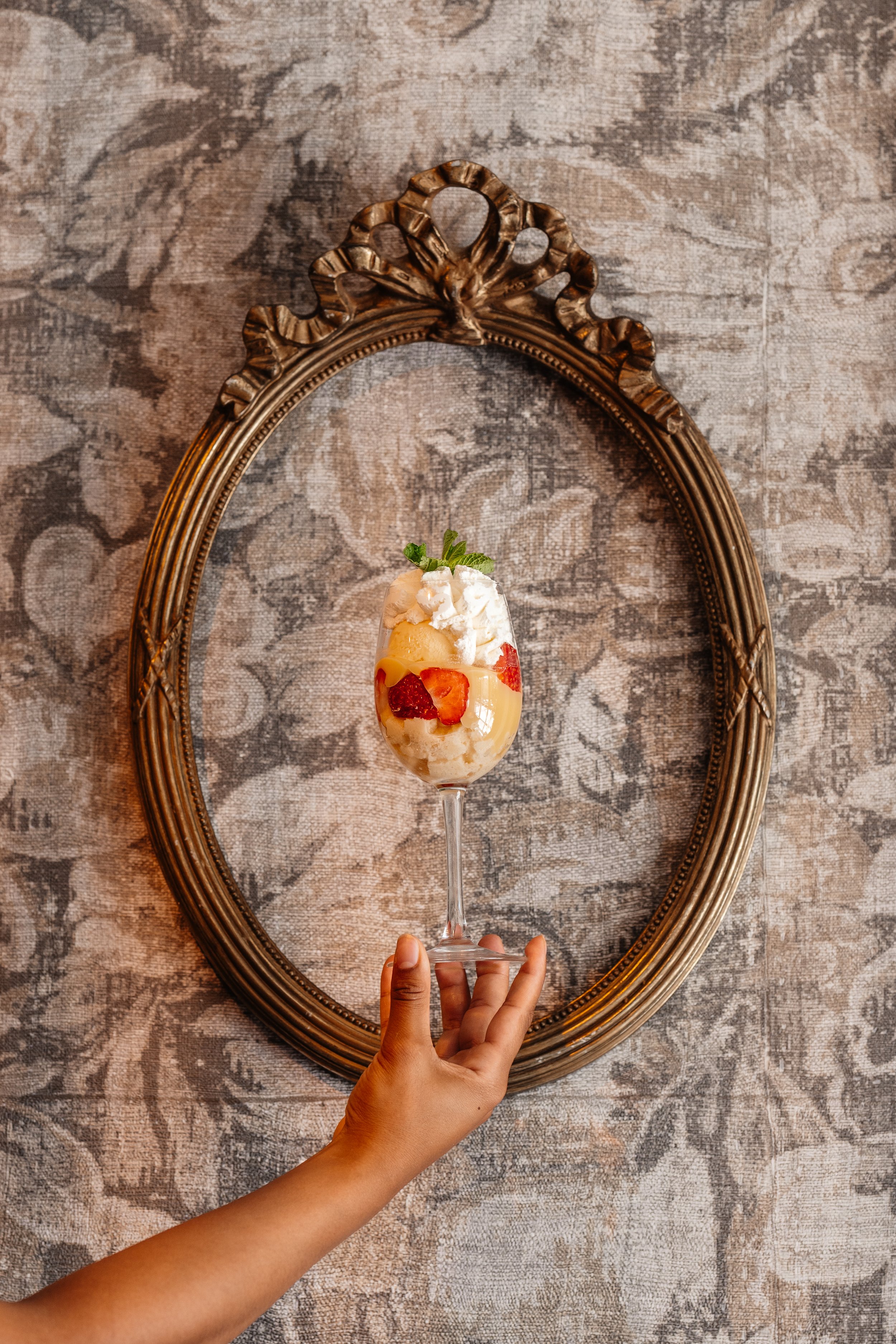 A hand holding a dessert glass filled with strawberries, pineapple, whipped cream, and garnished with mint, positioned in front of an ornate, oval-shaped mirror on a patterned wall.