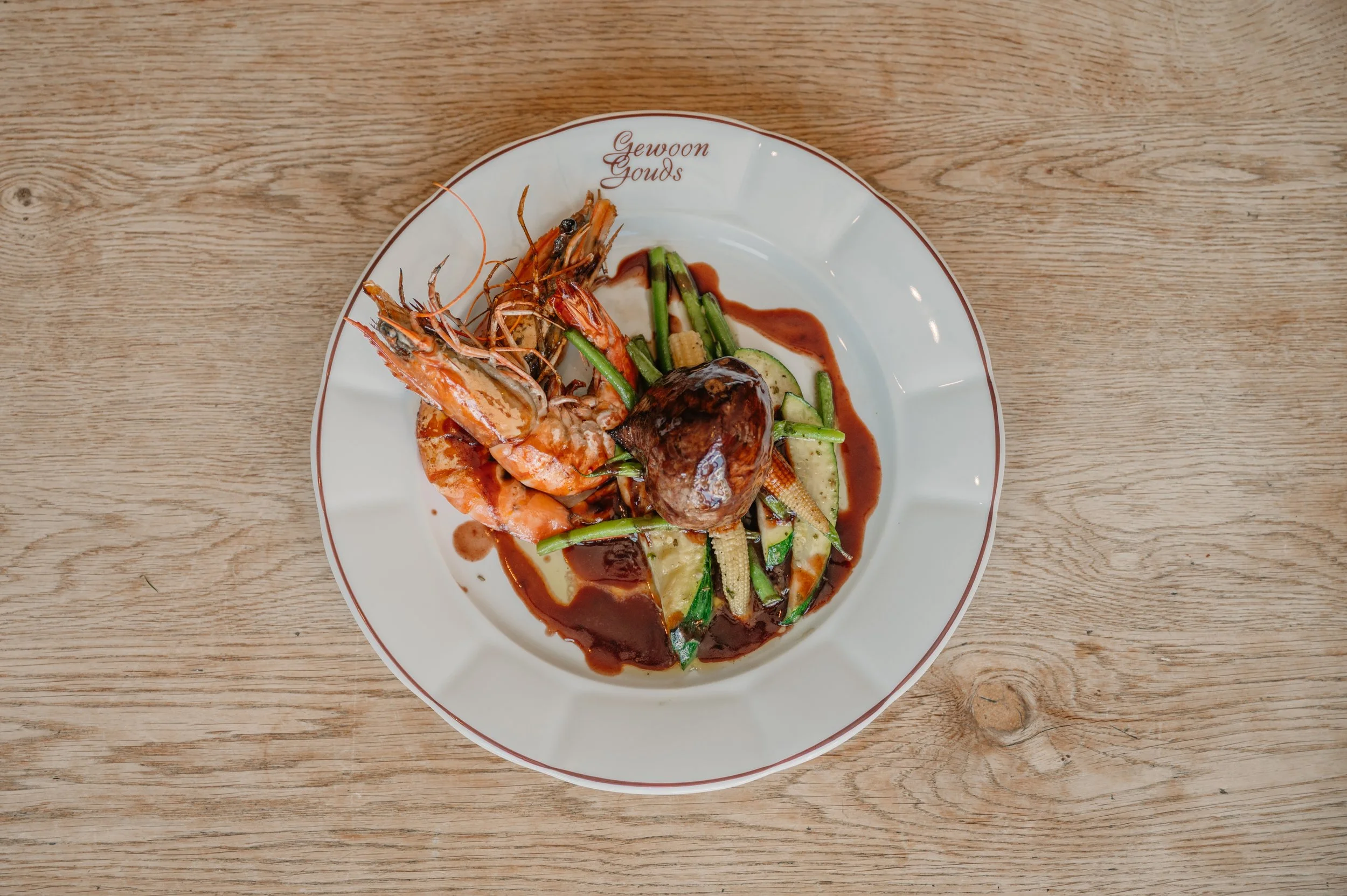 Seafood dish with shrimp, green vegetables, baby corn, zucchini, and brown sauce on a white plate with red rim, on a wooden table.