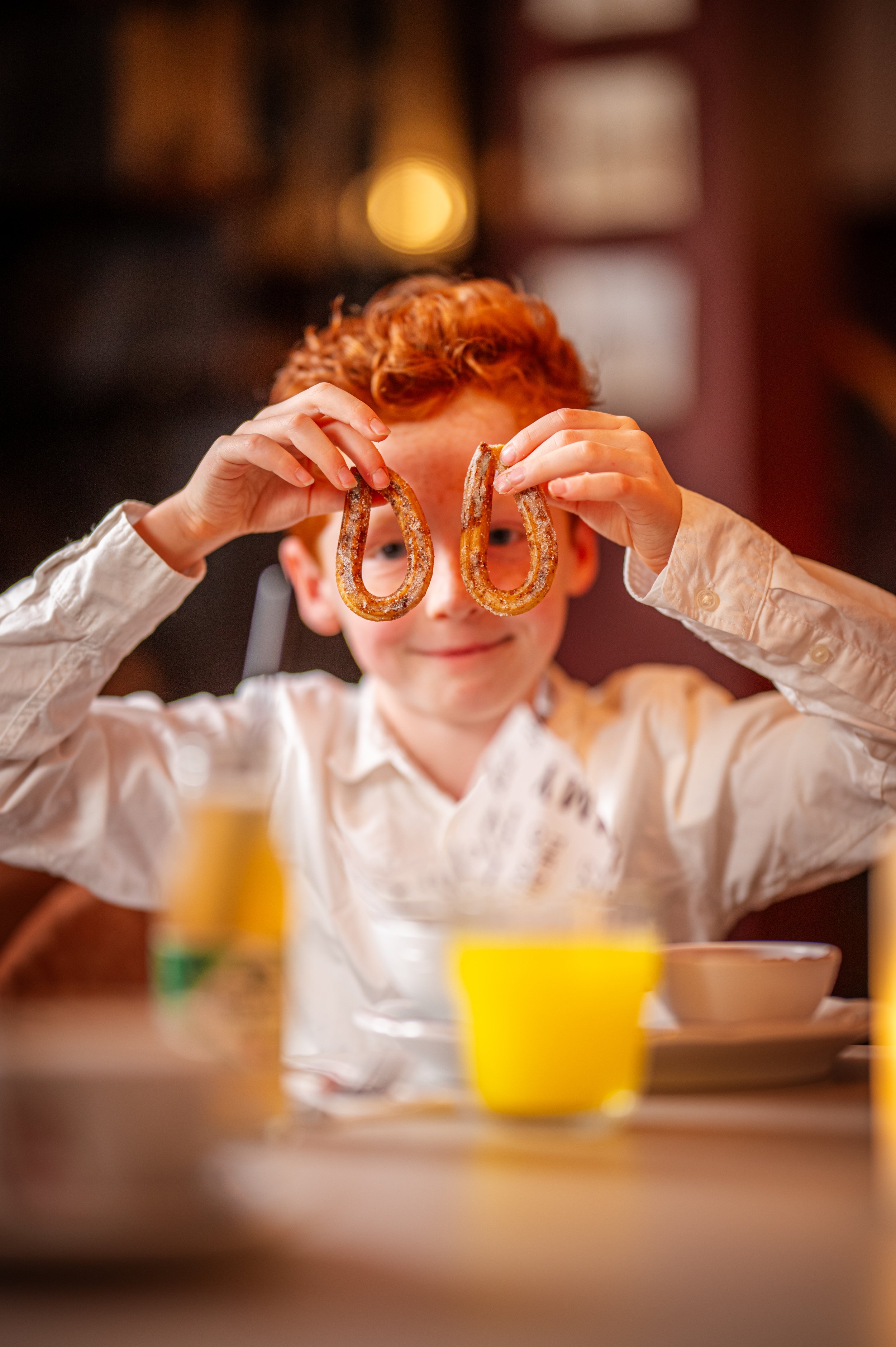A young boy with red hair holding two churros in front of his face in a restaurant.