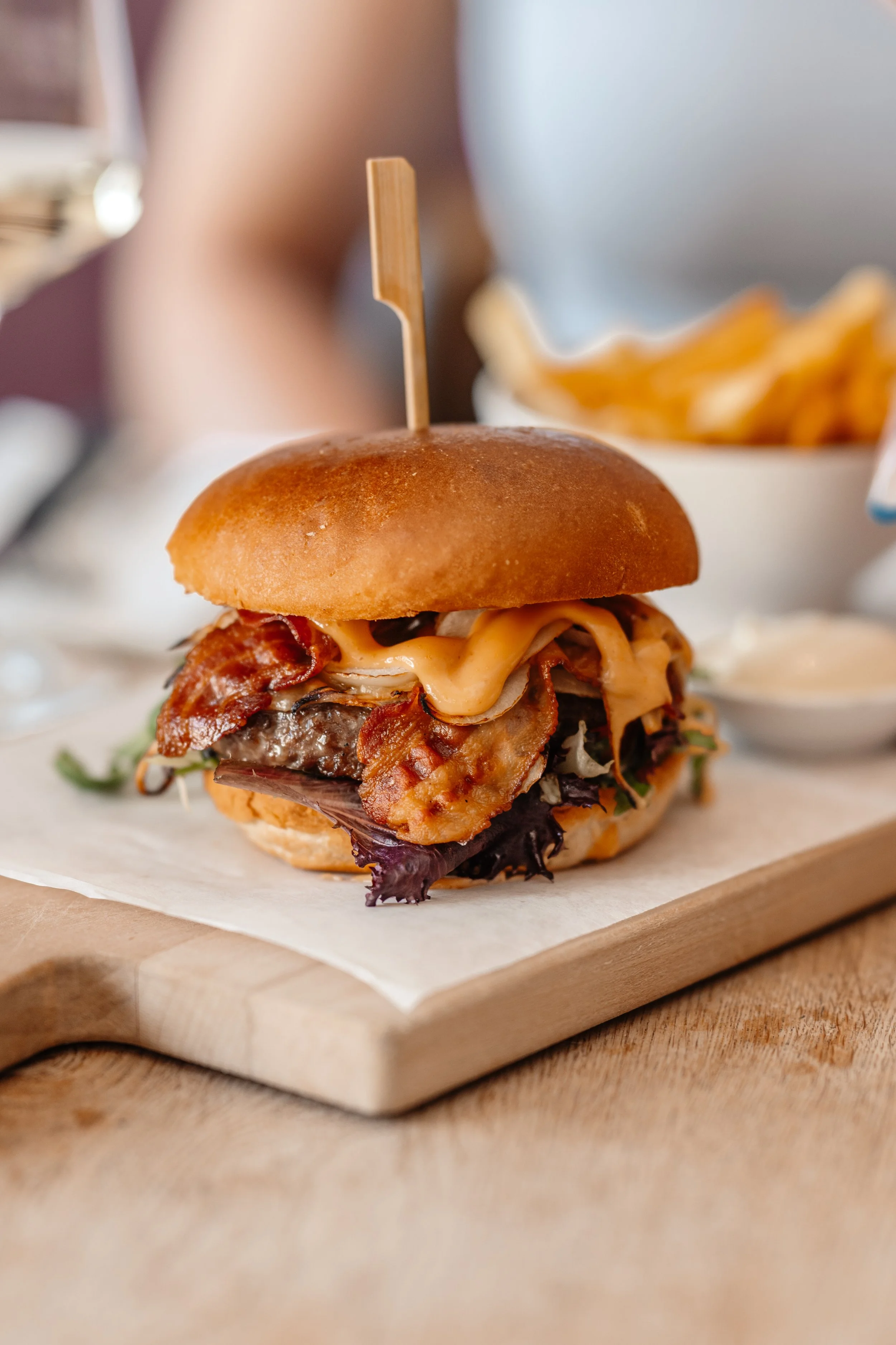 A close-up of a cheeseburger with bacon, lettuce, and cheese in a bun, served on a wooden board with a side of French fries in the background.