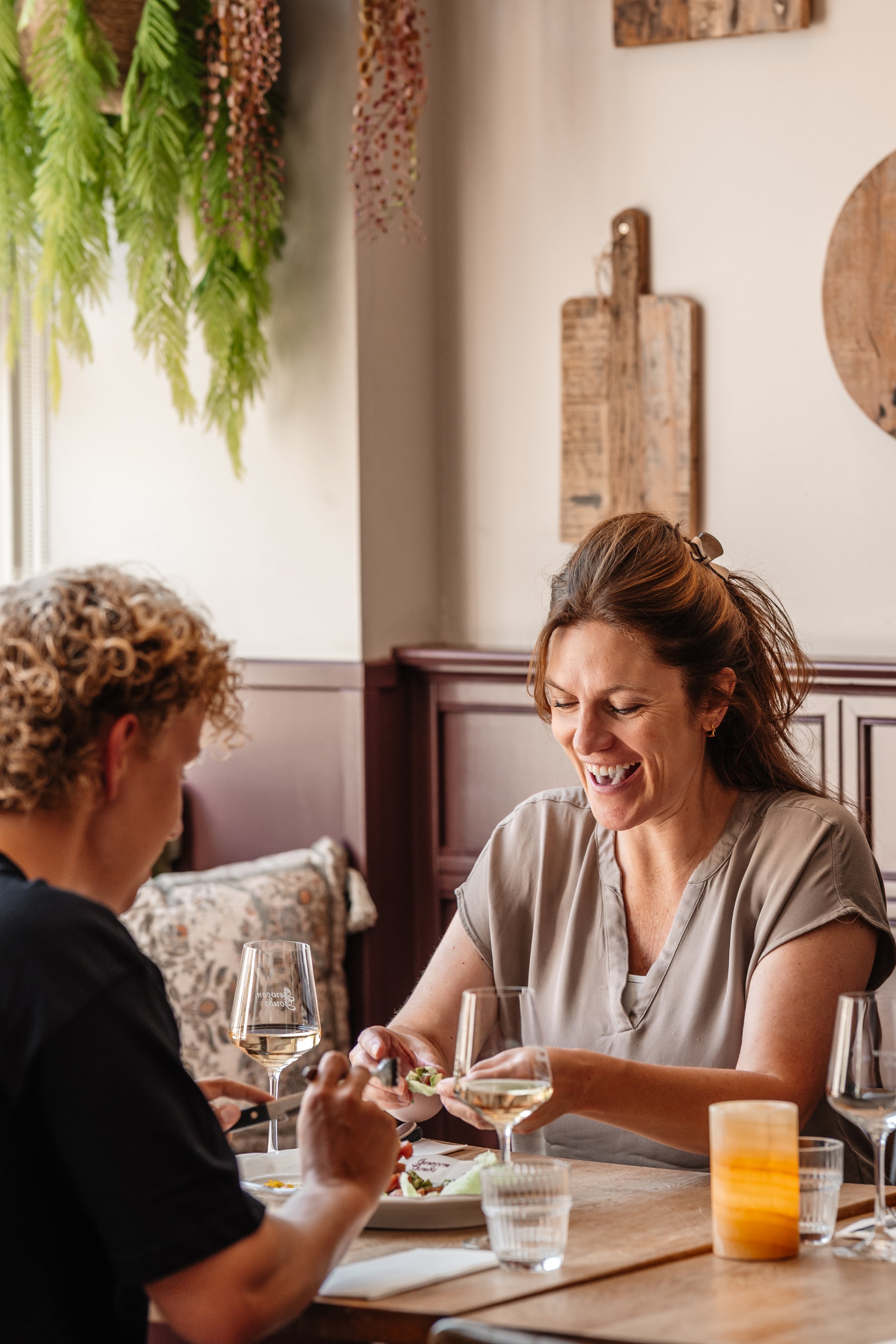Two women laughing and enjoying a meal with glasses of white wine on a wooden table in a cozy restaurant.
