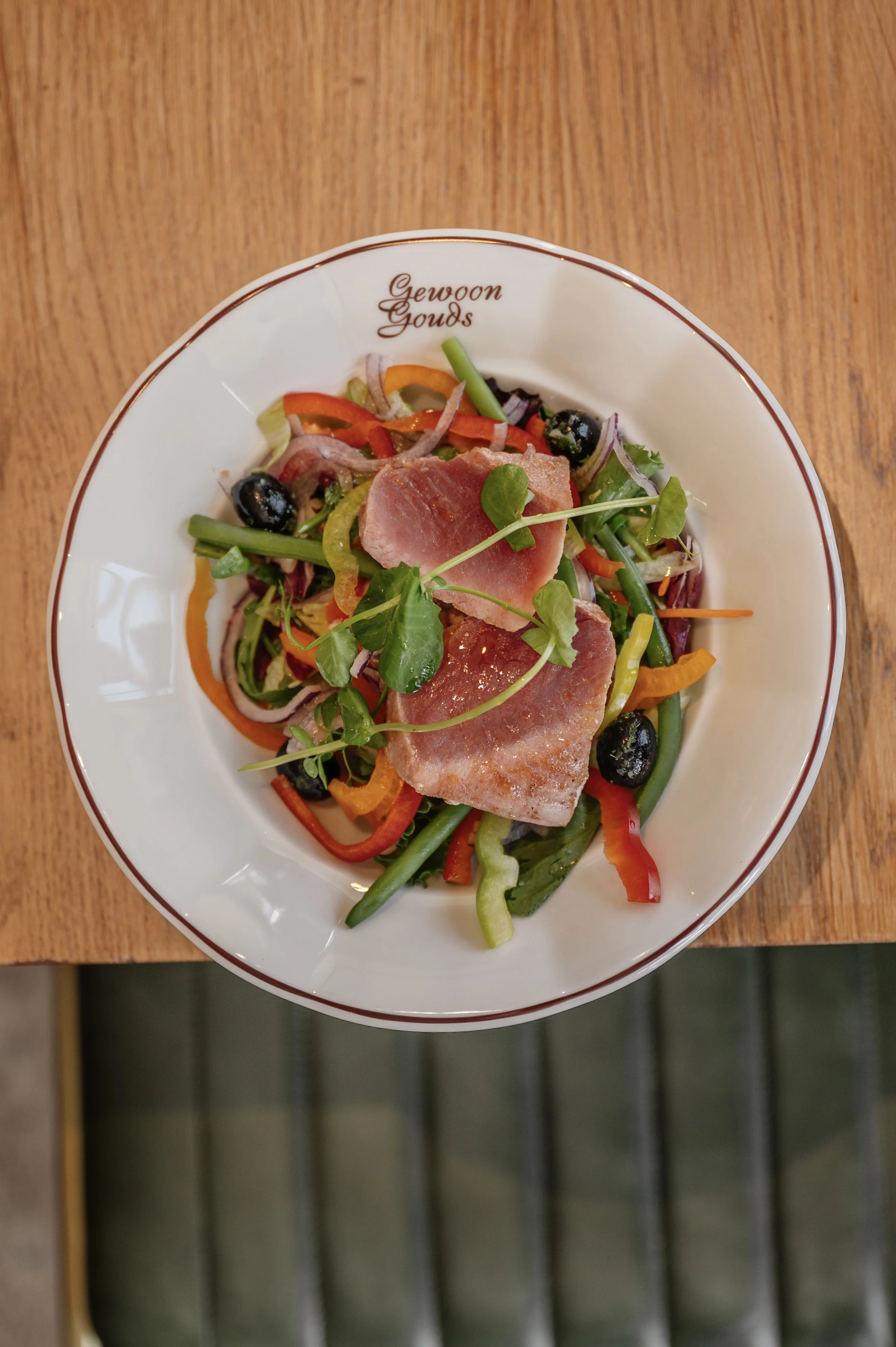 Salad with greens, sliced ham, olives, red and yellow bell peppers, and microgreens in a white bowl on a wooden table.